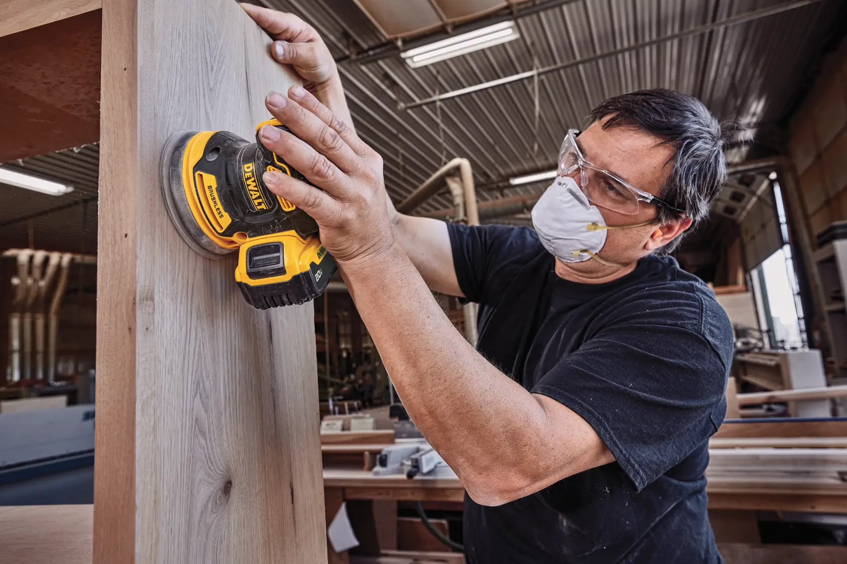 A person wearing a dust mask is using a DEWALT cordless sander (model DCW210B) to sand a wooden surface in a workshop.