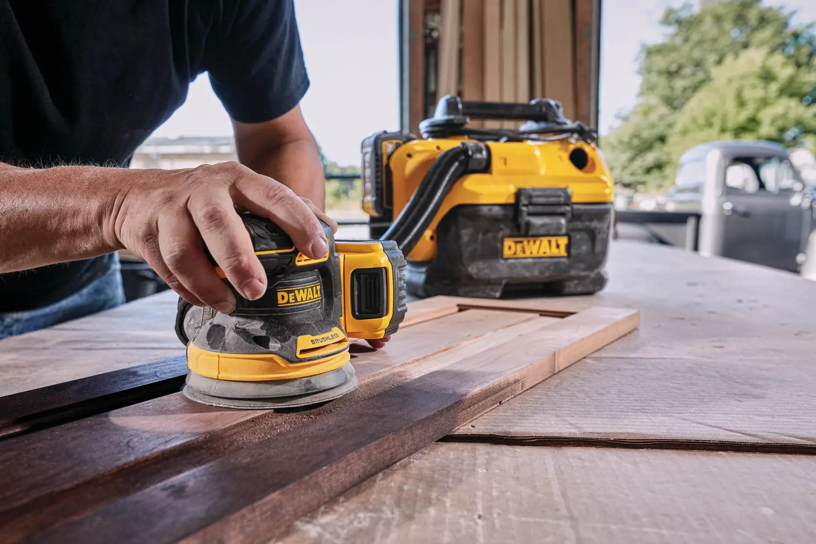 A person uses a DEWALT cordless orbital sander to smooth a piece of wood on a workbench, with a DEWALT vacuum system visible in the background.