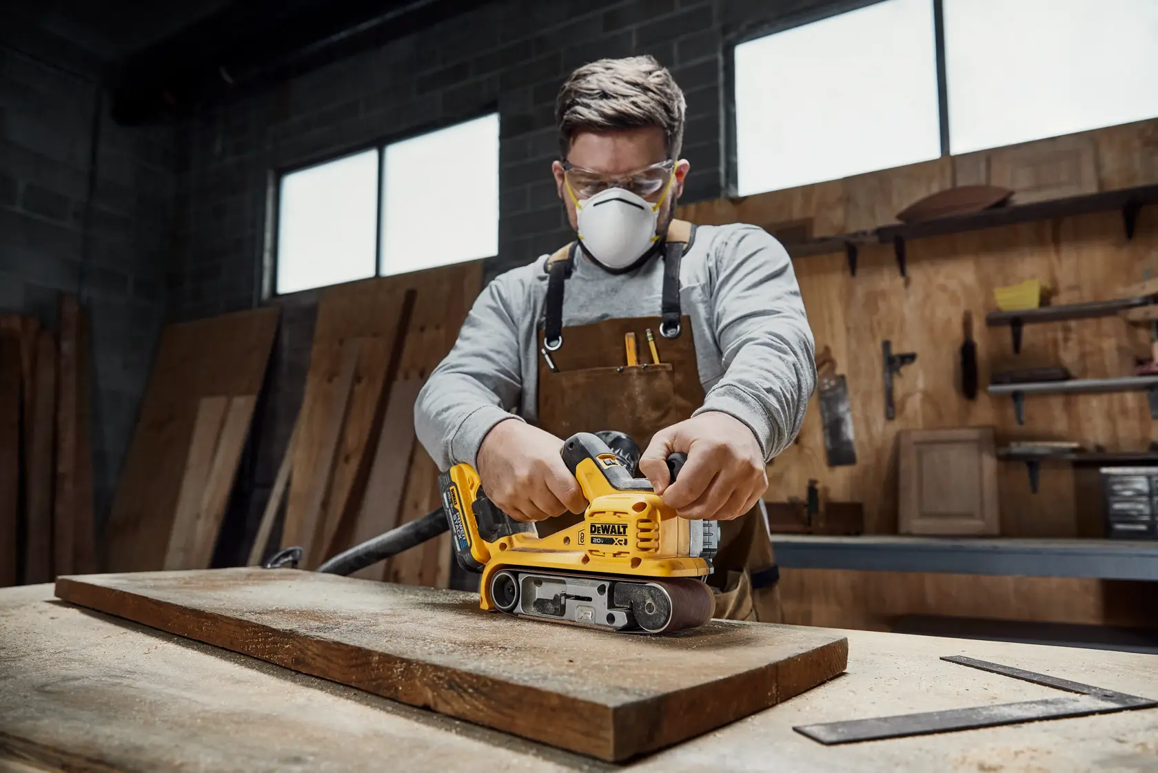 A person using a DEWALT DCW220B cordless belt sander on a wooden plank in a workshop. The person's face is blurred for privacy.