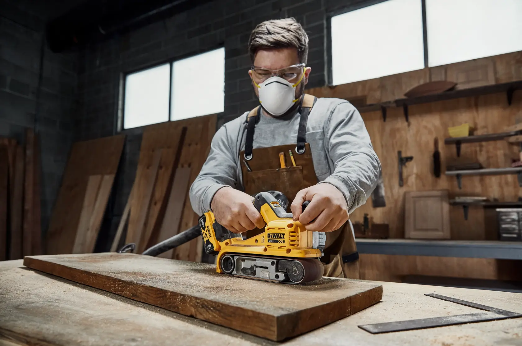 A person wearing a brown apron is using a yellow DEWALT DCW220B belt sander on a wooden plank in a workshop. The person's face is blurred for privacy. Various wooden boards and workshop tools are visible in the background.