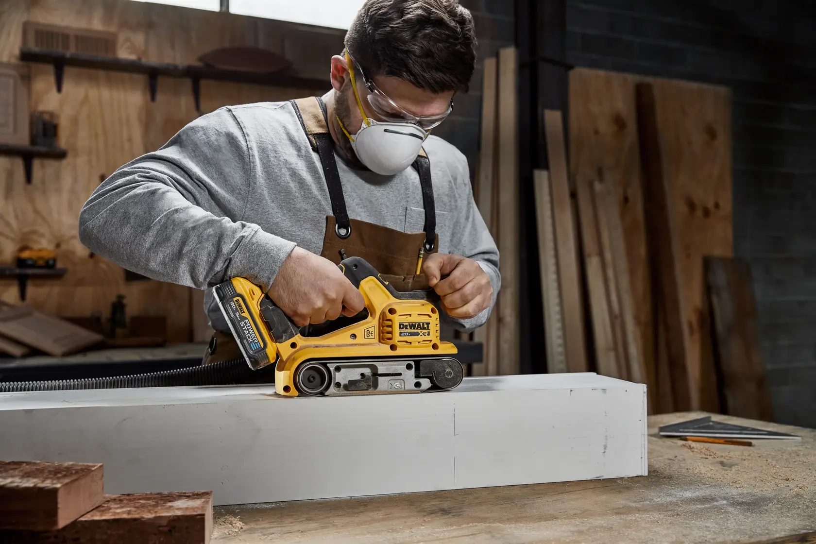 A person using a DEWALT DCW220B cordless belt sander on a wooden plank in a workshop. The person's face is blurred for privacy.