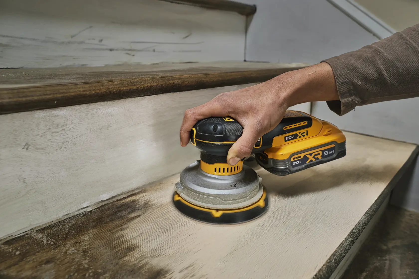 Une personne utilise une ponceuse orbitale aléatoire sans fil DEWALT pour poncer un escalier en bois.