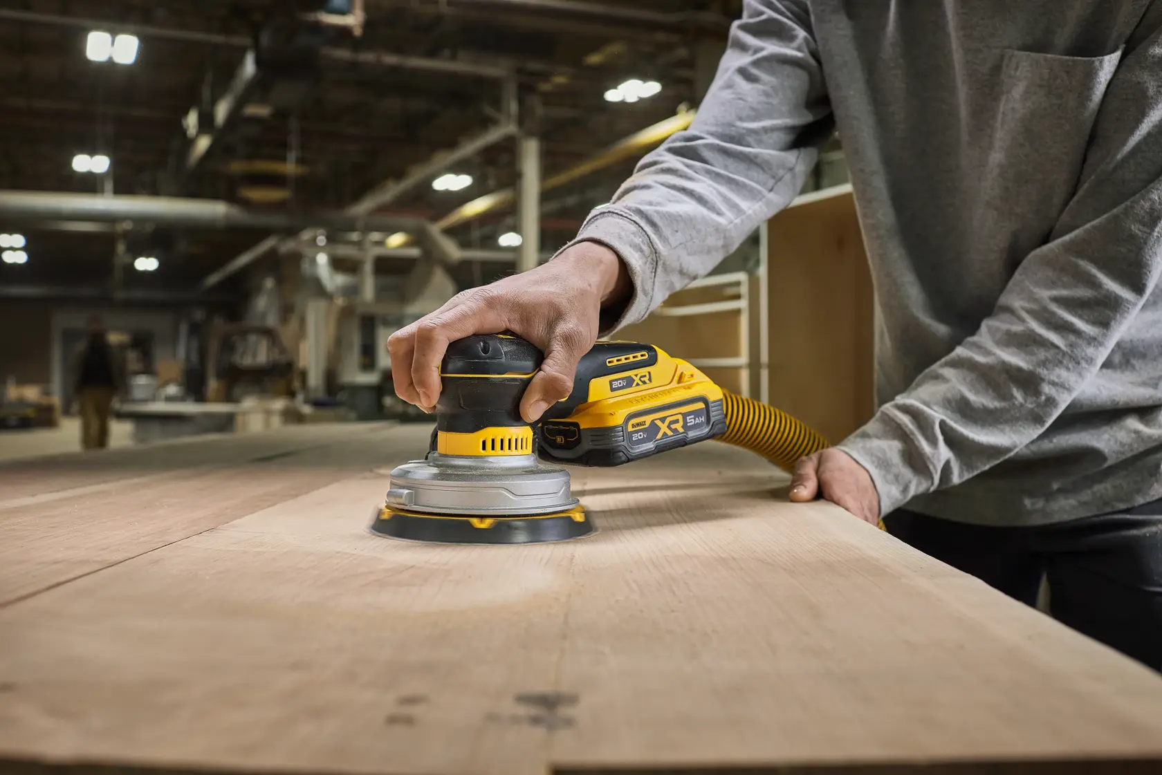 Une personne utilise une ponceuse sans fil DEWALT DCW230B avec un tuyau d'extraction de poussière pour lisser une grande surface en bois dans un atelier.