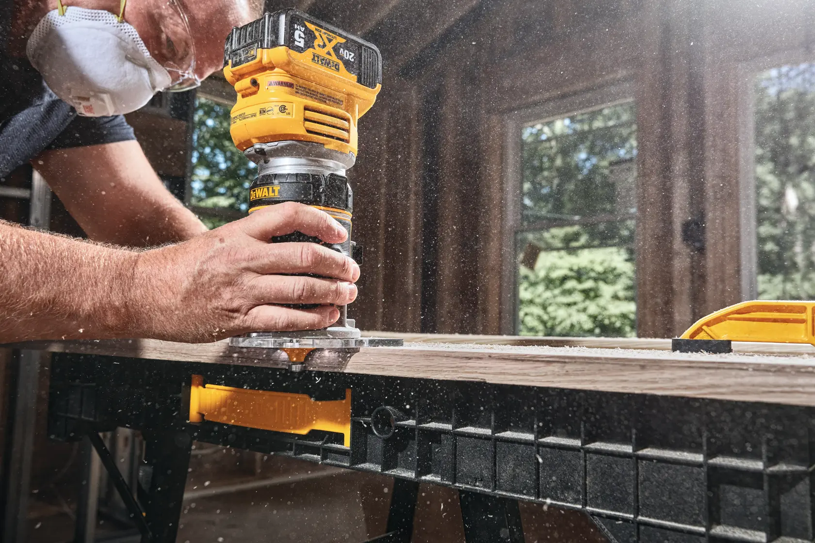Close-up of a person using a DEWALT cordless router to shape the edge of a wooden board on a workbench, with sawdust flying and the person wearing a protective mask.