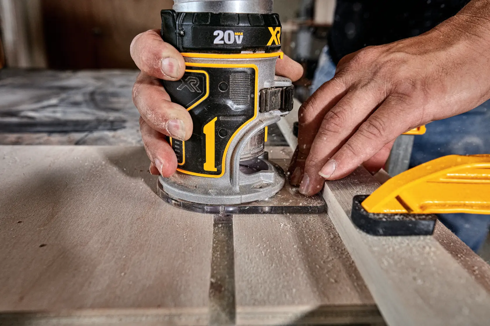 Close-up of hands using a DEWALT cordless router tool on a piece of wood, with a yellow clamp holding the wood in place.