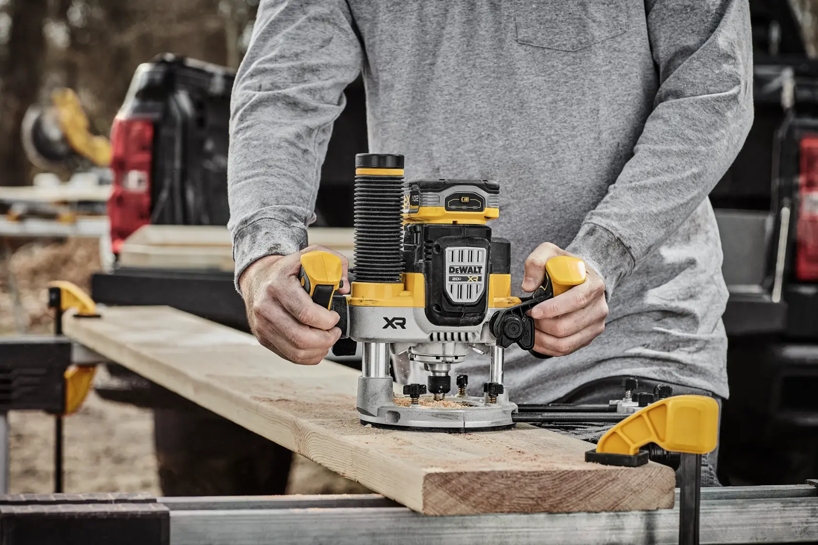 A person operates a DEWALT cordless wood router on a plank of wood, which is securely clamped to a workbench outdoors. The router is yellow and black, displaying the DEWALT branding.