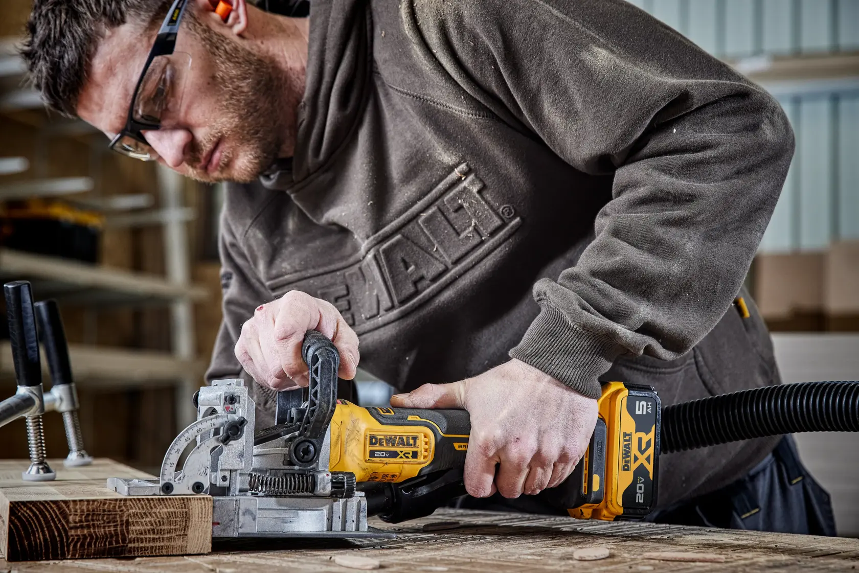 A person using a DEWALT DCW682B cordless woodworking tool with an attached vacuum hose on a wooden workbench. The individual is wearing a DEWALT branded sweatshirt and working with clamps and wood pieces in a workshop environment.