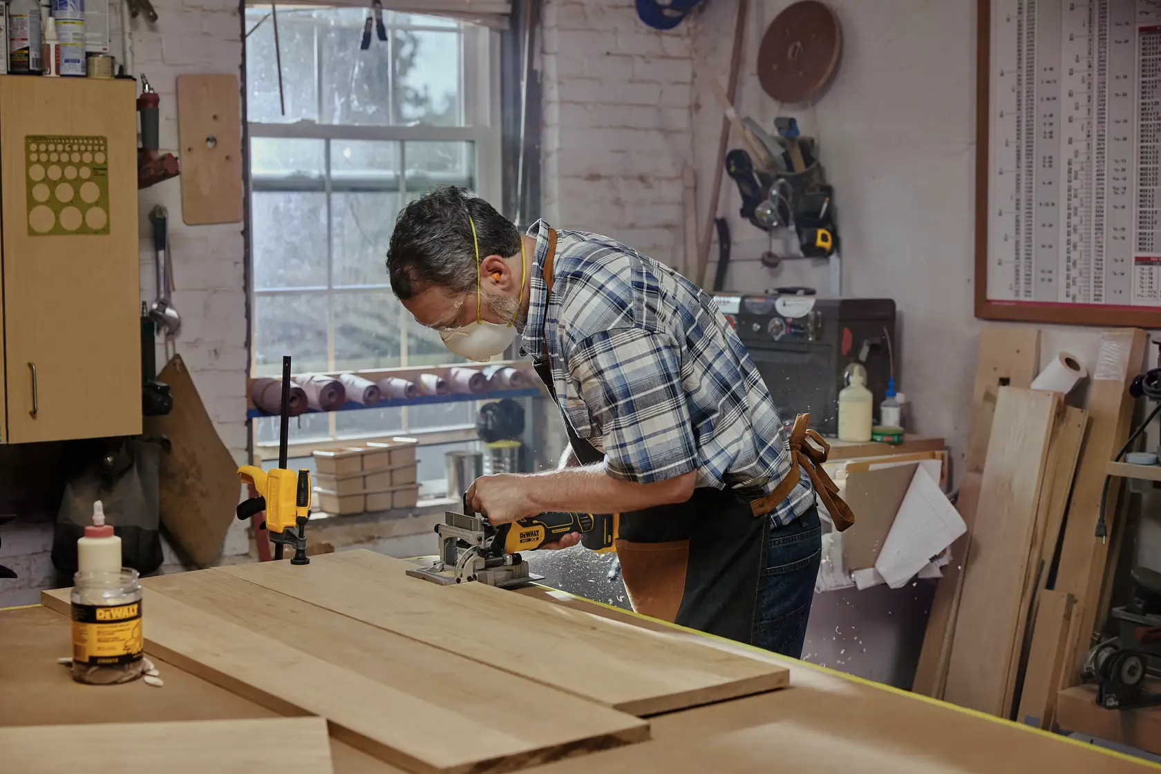 A person is using a DEWALT cordless biscuit joiner tool to cut slots into wooden boards in a workshop. Various woodworking materials and tools are visible around the workspace, including clamps and a bottle of glue.