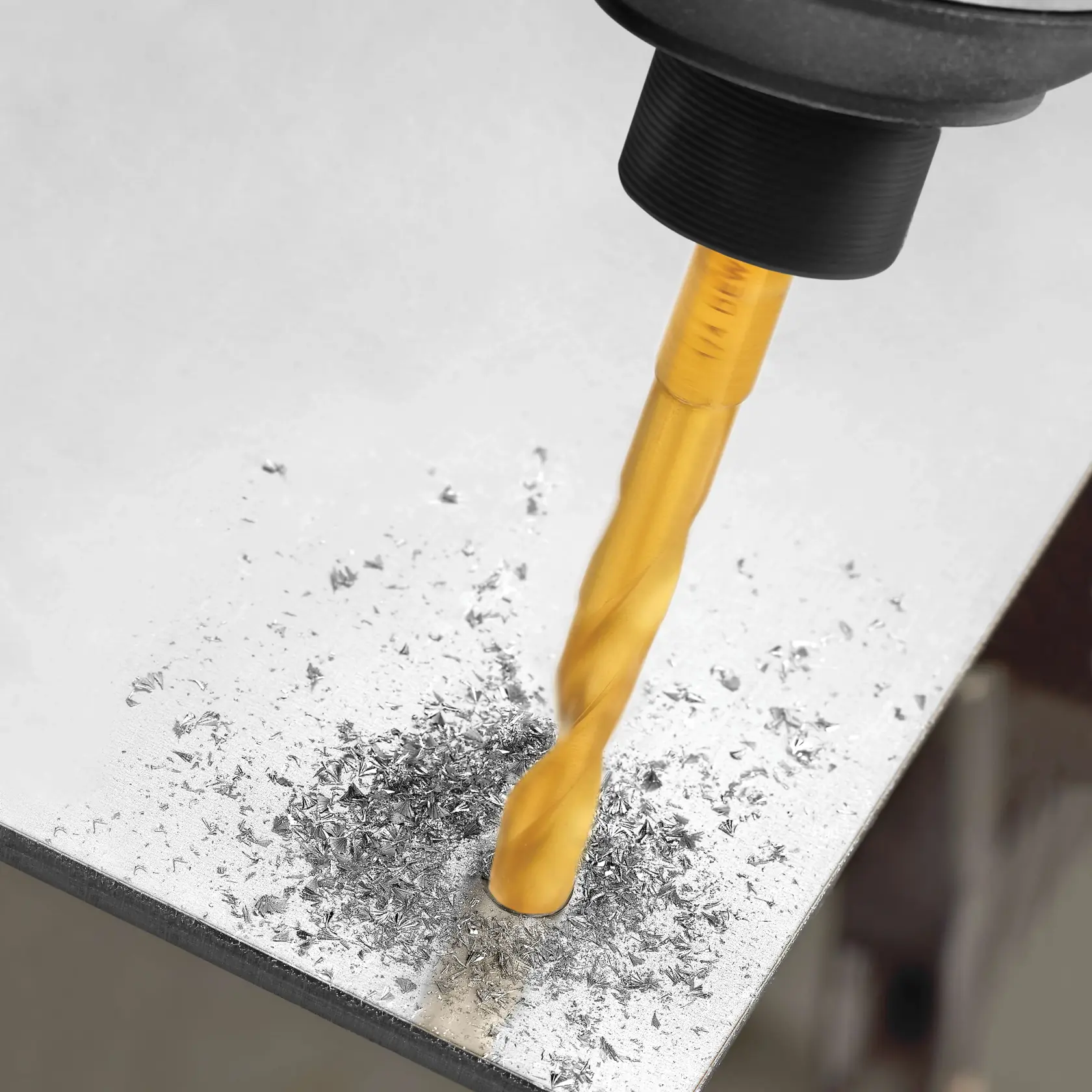 A gold-colored drill bit is being used to drill into a metal surface, creating metal shavings around the hole.