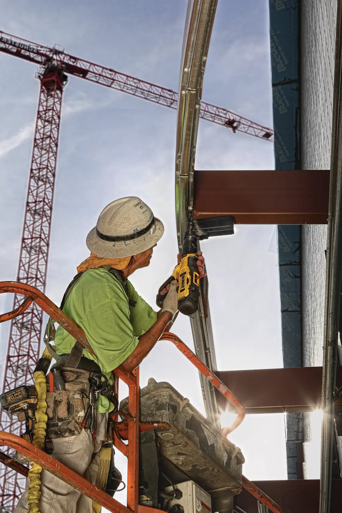 A construction worker wearing safety gear stands on an elevated platform and uses a DEWALT power tool to fasten metal framing on a building. Tall red cranes are visible in the background against a blue sky.