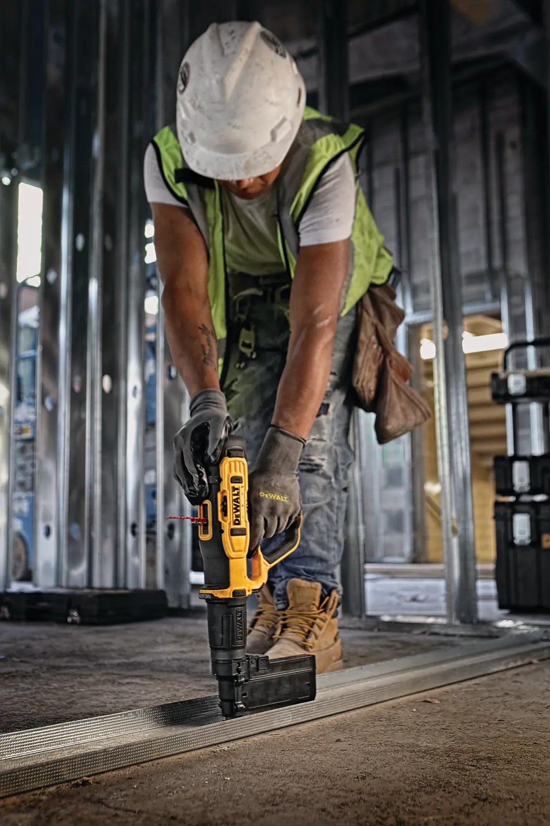 A construction worker wearing a safety vest, gloves, and hard hat uses a DEWALT tool to secure metal framing to the floor inside a building under construction.