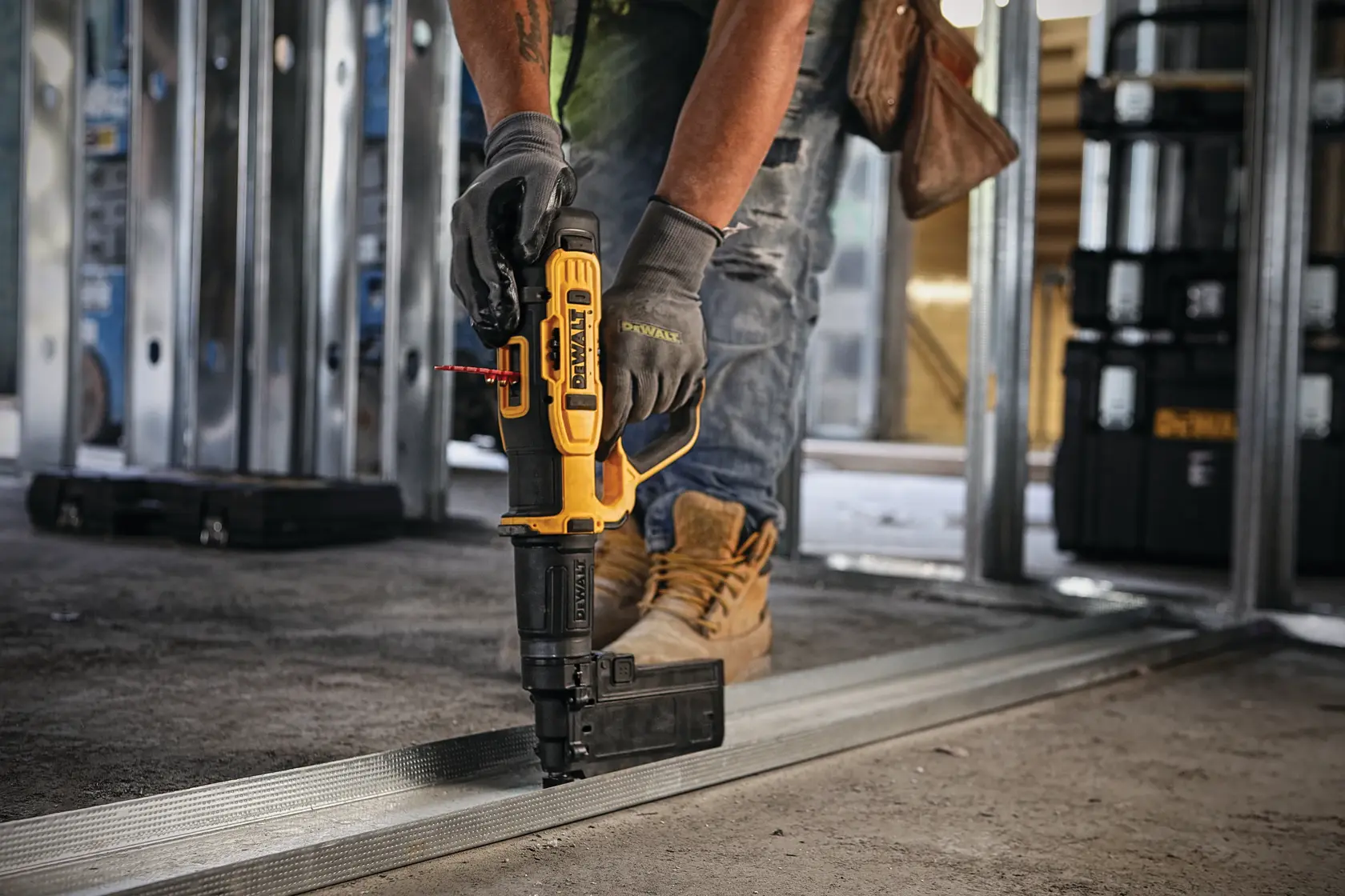 A worker wearing gloves and boots uses a DEWALT power tool to fasten metal framing to the floor at a construction site. The tool is yellow and black, and the worker is surrounded by metal studs and toolboxes.