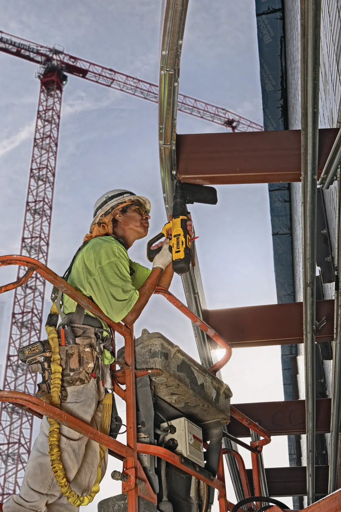 A construction worker wearing safety gear uses a DEWALT tool to fasten metal framing while standing on an elevated platform. A red crane and steel beams are visible in the background.