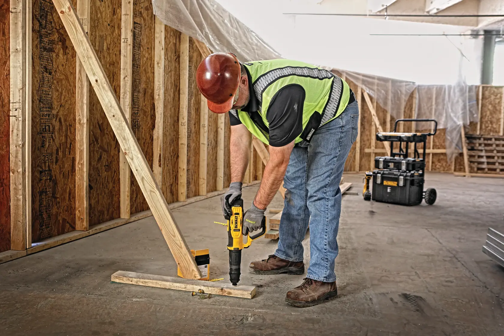 A construction worker wearing a safety vest and hard hat uses a DEWALT power tool to fasten a wooden beam to a wooden floor brace in an unfinished building. A DEWALT tool box is visible in the background.