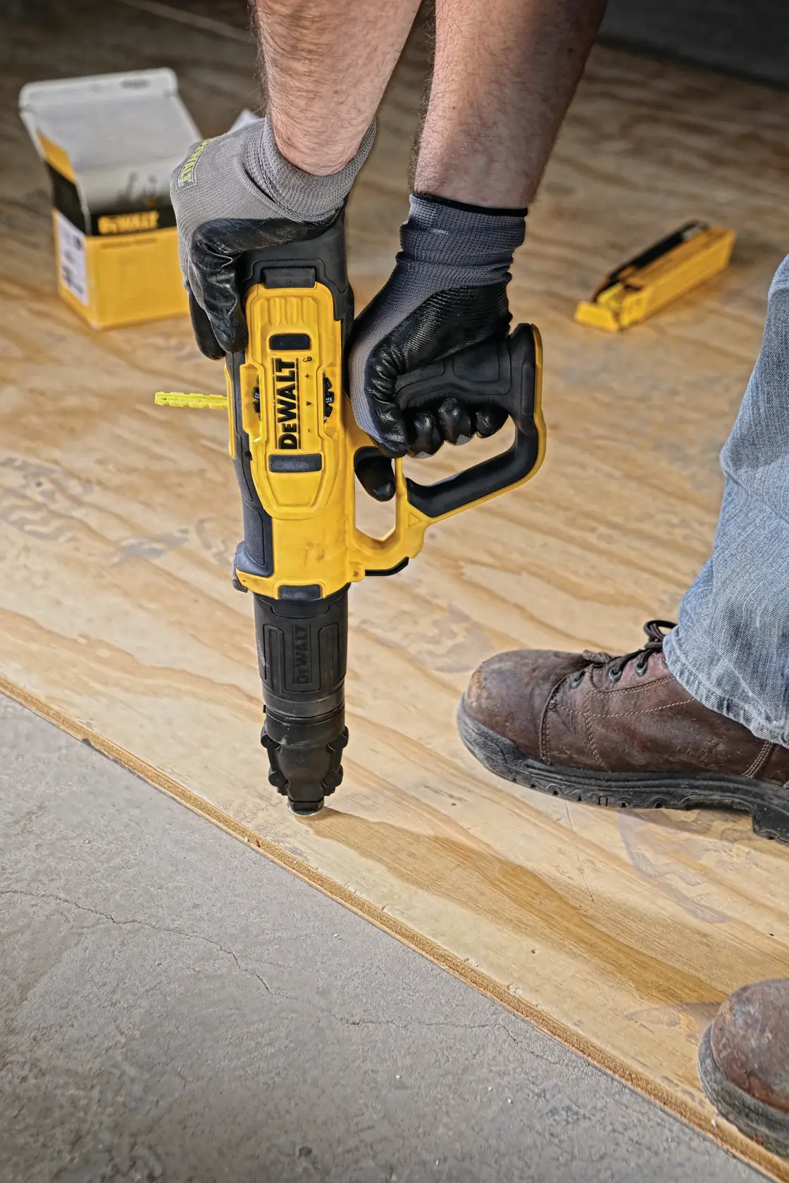 Person using a DEWALT yellow and black tool to secure plywood flooring, wearing gloves and work boots. Other tools and a DEWALT box are visible in the background.