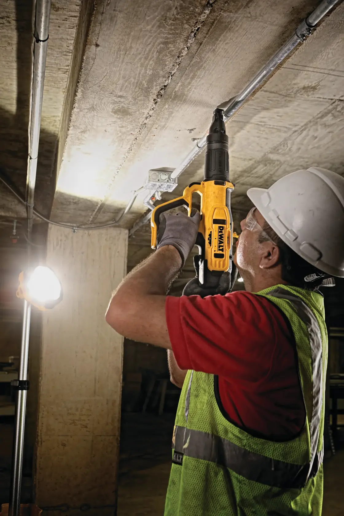 A person wearing a safety vest, gloves, and a hard hat is using a yellow DEWALT powder-actuated tool to fasten fixtures to a concrete ceiling in an industrial or construction setting.