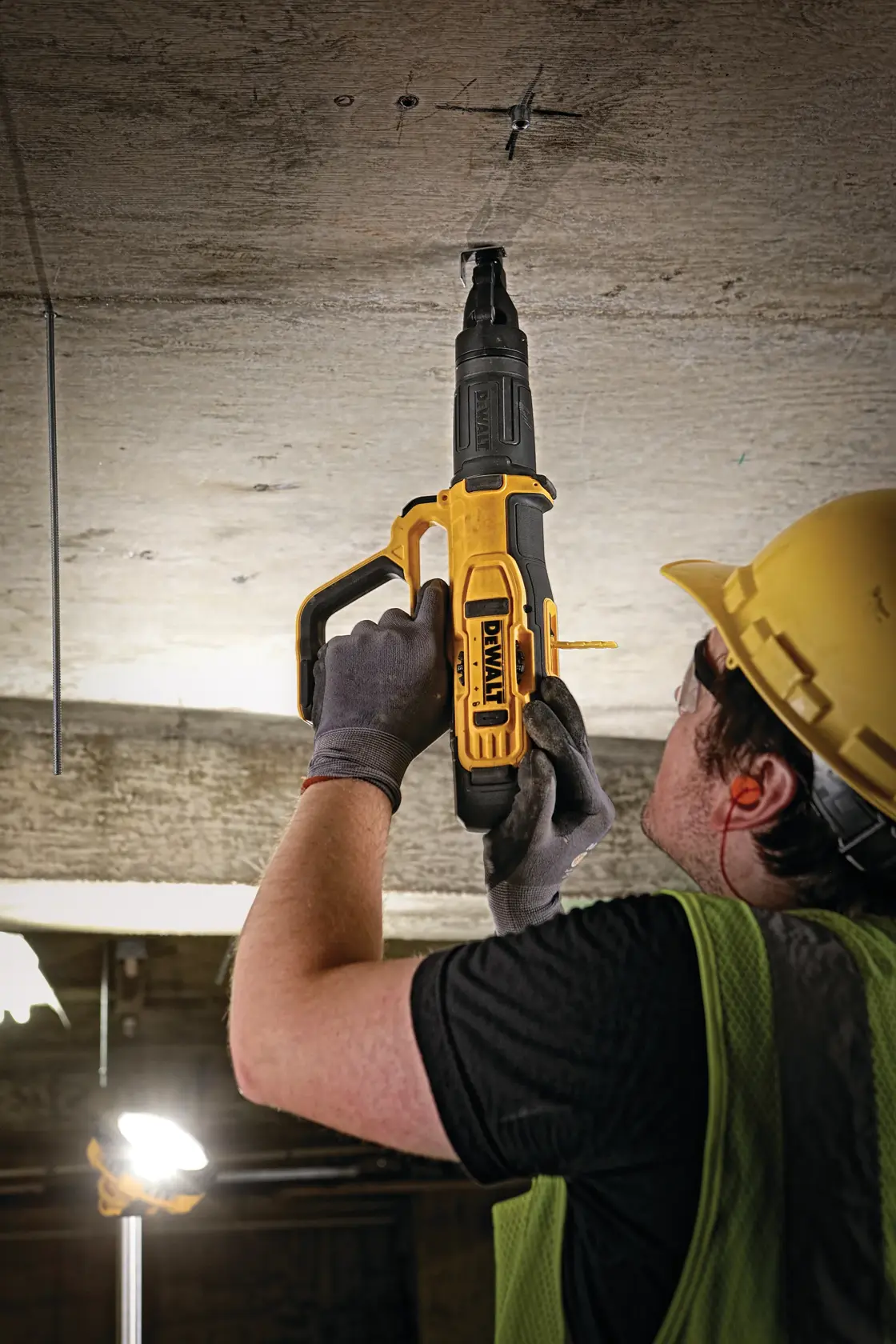 A worker wearing a yellow safety helmet, gloves, and a green safety vest is using a DEWALT powder-actuated tool to fasten a fixture into a concrete ceiling on a construction site.