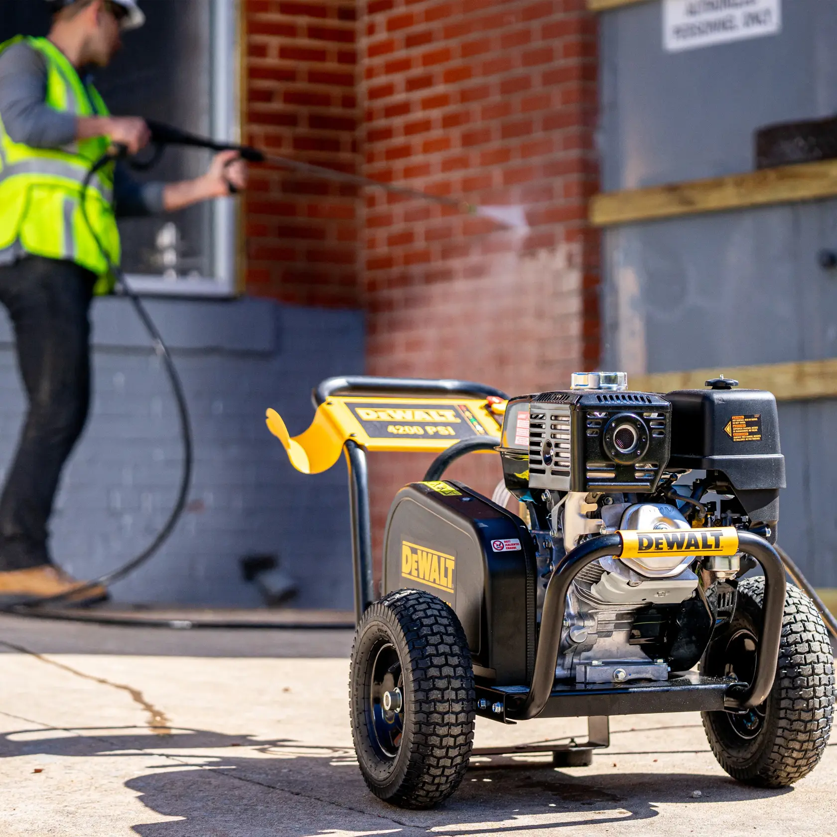 A DEWALT pressure washer, model DH4240B, is positioned outdoors on a paved surface. The pressure washer features large wheels and a sturdy frame. In the background, a person wearing a safety vest uses the pressure washer to clean a building exterior with brick and gray walls.