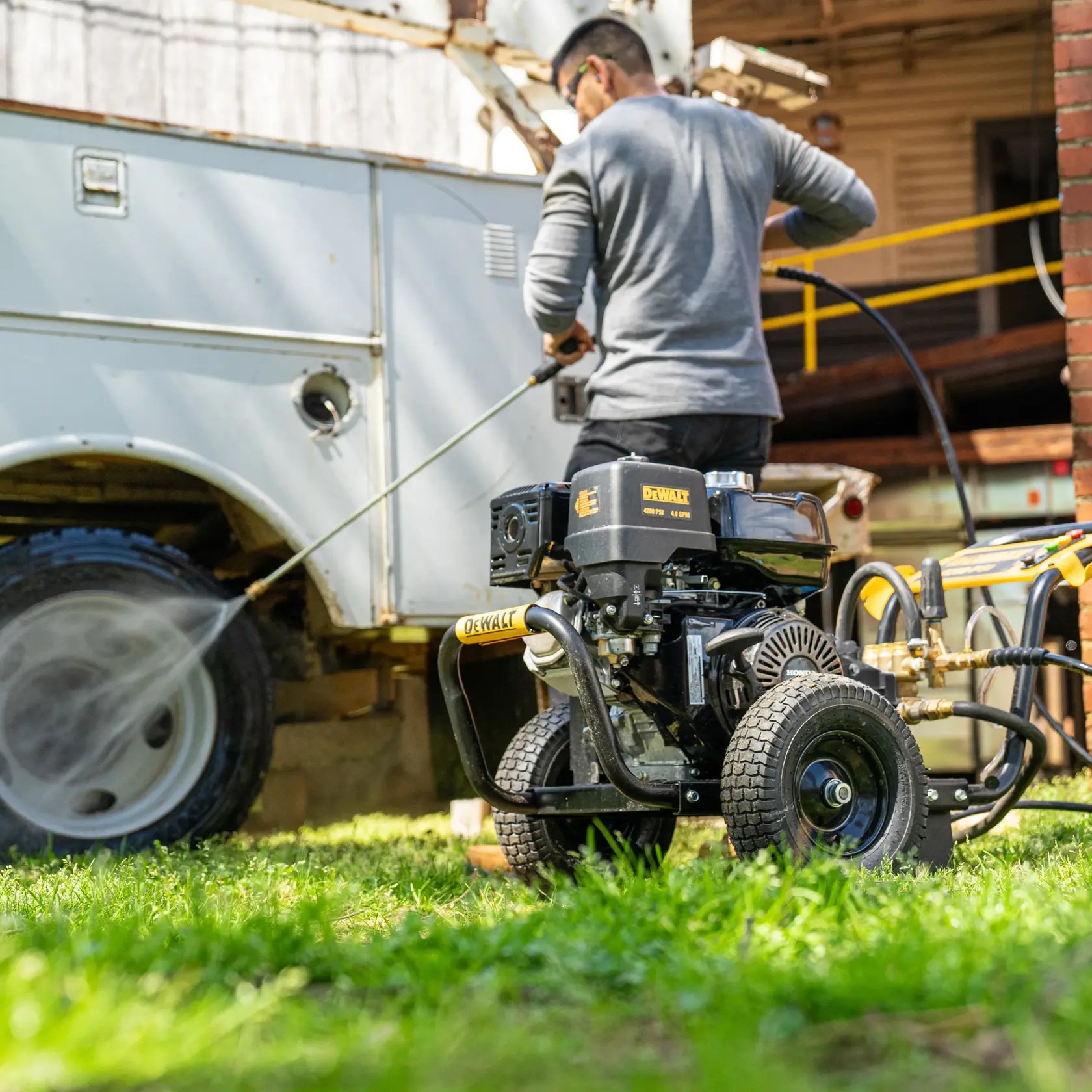 A person uses a DEWALT pressure washer (model DH4240B) to clean the wheel of a utility vehicle outdoors. The pressure washer is placed on grass and is connected with hoses, spraying water onto the vehicle.