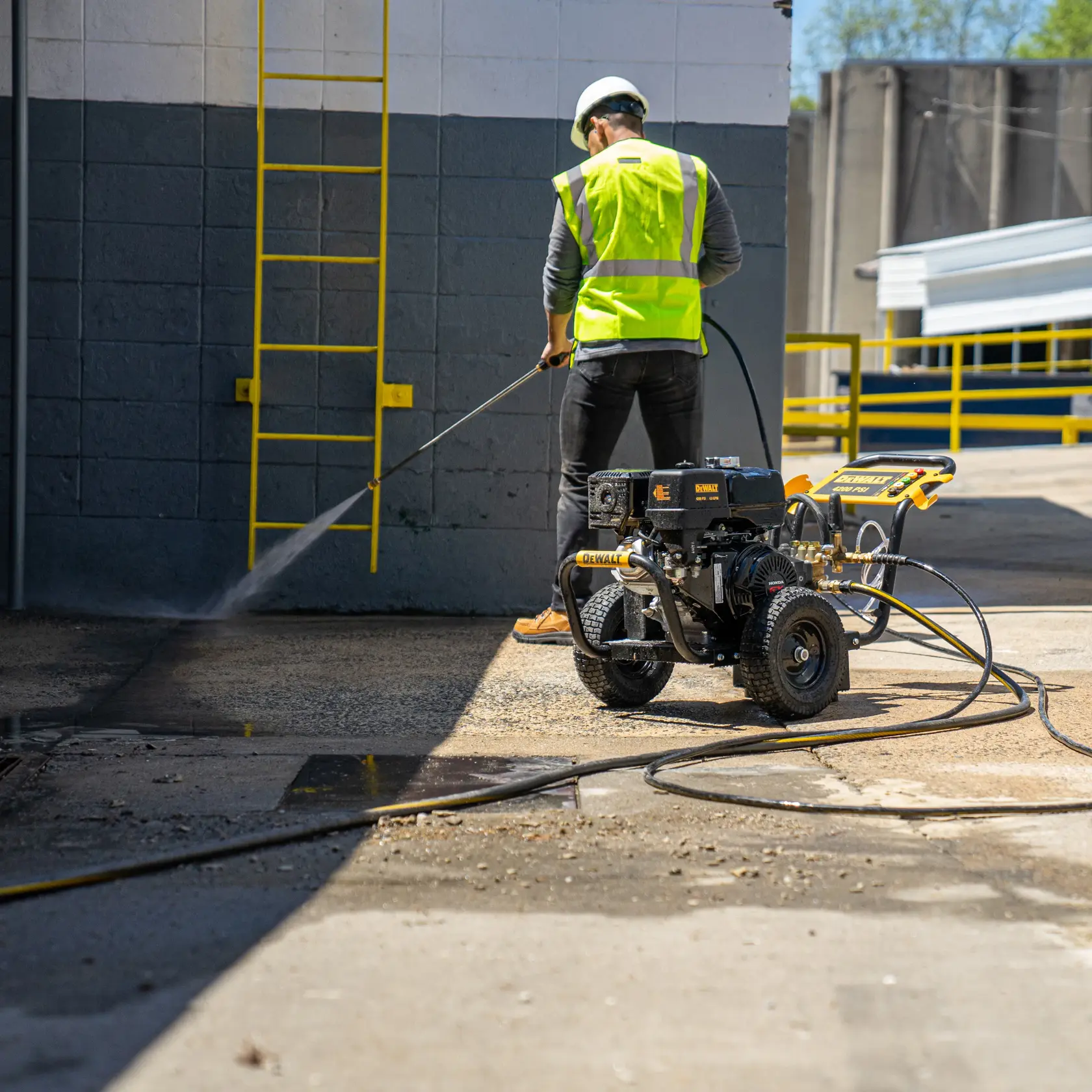 A person in safety gear operates a DEWALT pressure washer to clean a concrete surface outdoors near a building with a yellow ladder.