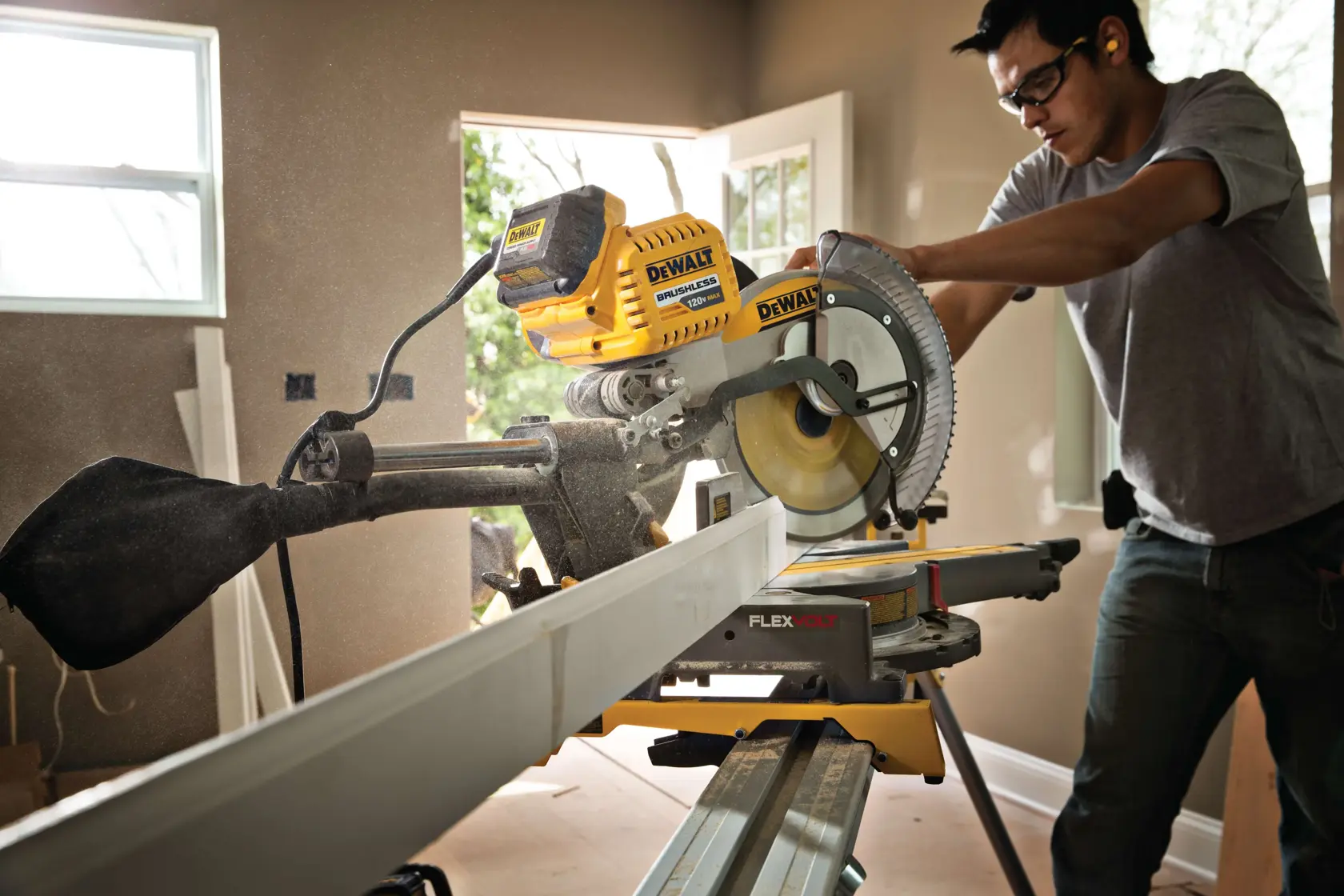 A person using a DEWALT DHS790AB FLEXVOLT miter saw to cut a long piece of wood indoors. The saw is yellow and black, and the worker is wearing ear protection. The room has natural light coming in through windows.