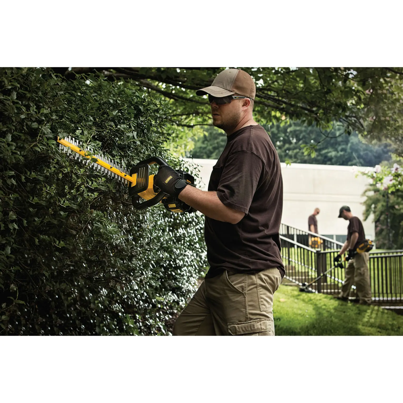 A person is using a DEWALT cordless hedge trimmer to trim bushes outdoors, wearing gloves and a cap. Two other people are also working in the background, using similar equipment.