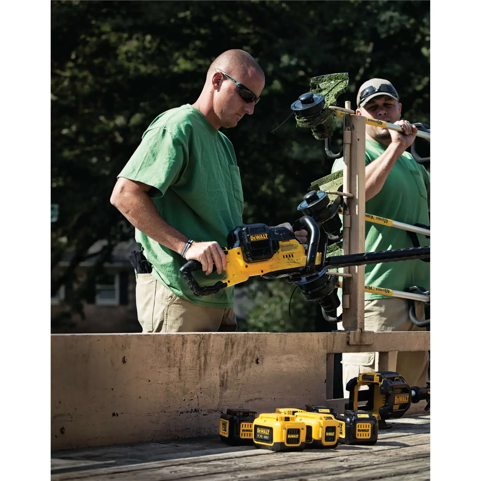 Two people with blurred faces are outdoors, working with DEWALT cordless tools. One is holding a yellow DEWALT chainsaw, while several DEWALT batteries and another tool are placed on a wooden surface. Both are wearing green shirts and beige pants.