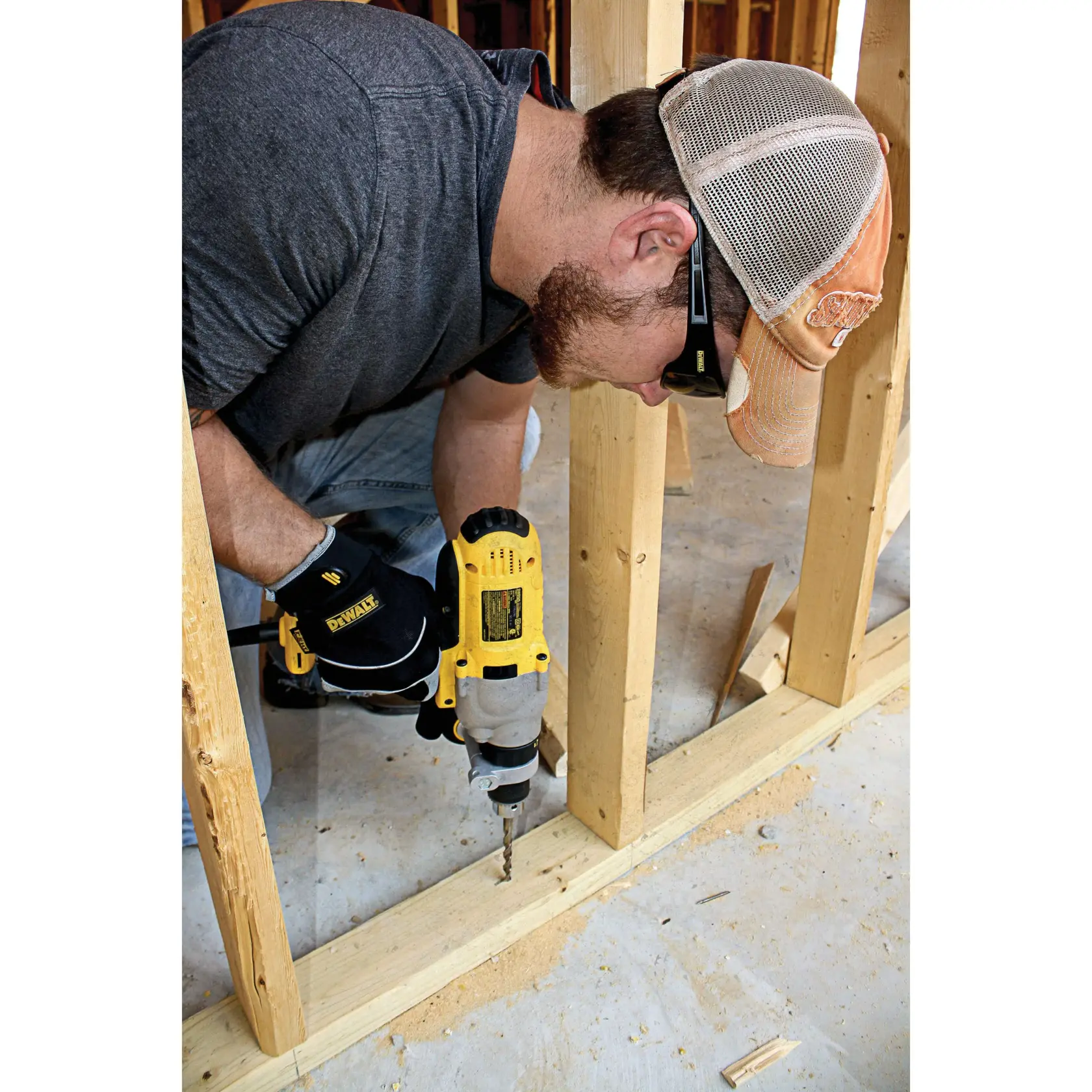 A person wearing a cap, safety glasses, and DEWALT branded gloves is using a yellow DEWALT power drill to drill into a wooden frame at a construction site.