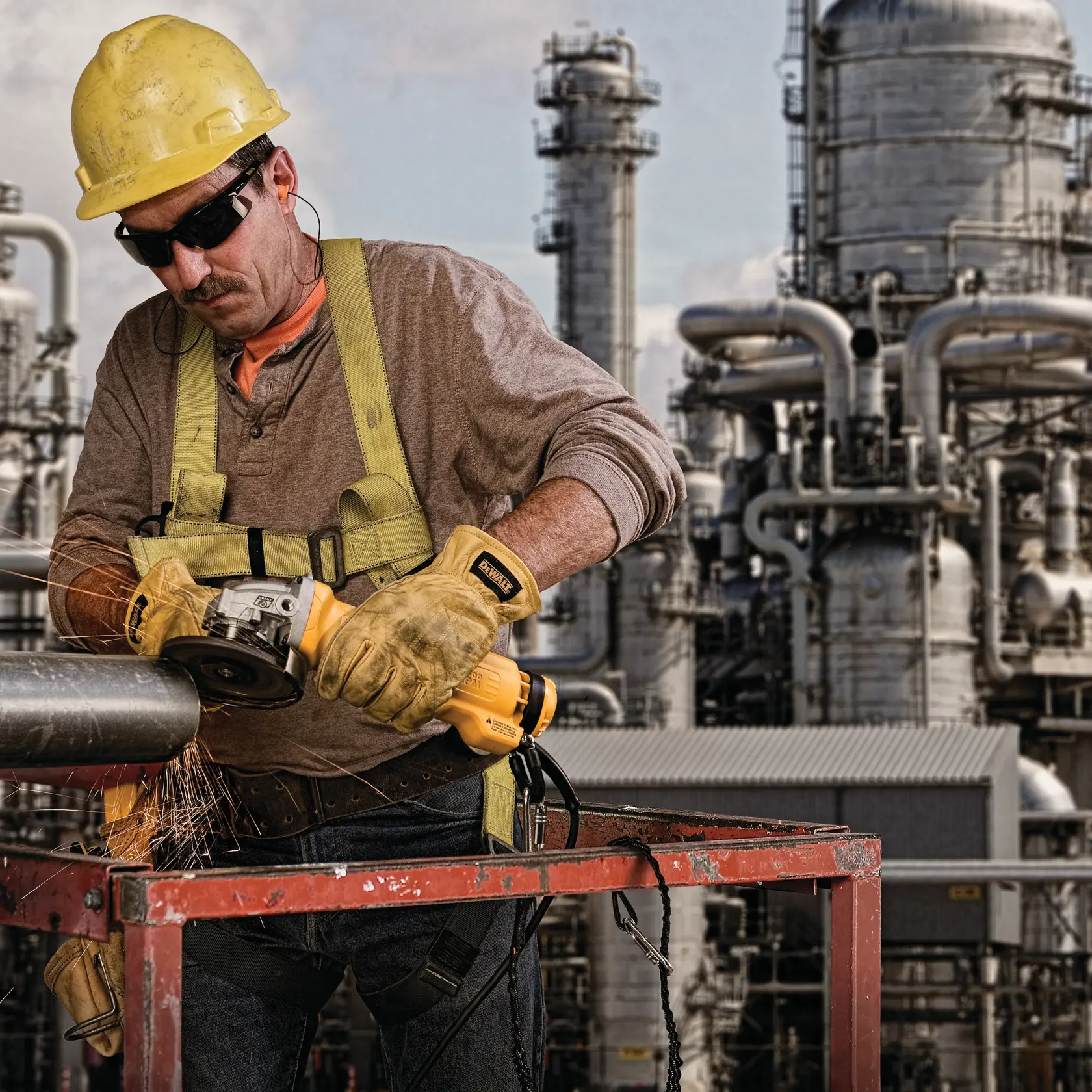 A worker wearing safety gear, including a hard hat, gloves, and harness, uses a yellow DEWALT power tool to cut a metal pipe, with sparks flying. In the background are large industrial structures with pipes and towers.