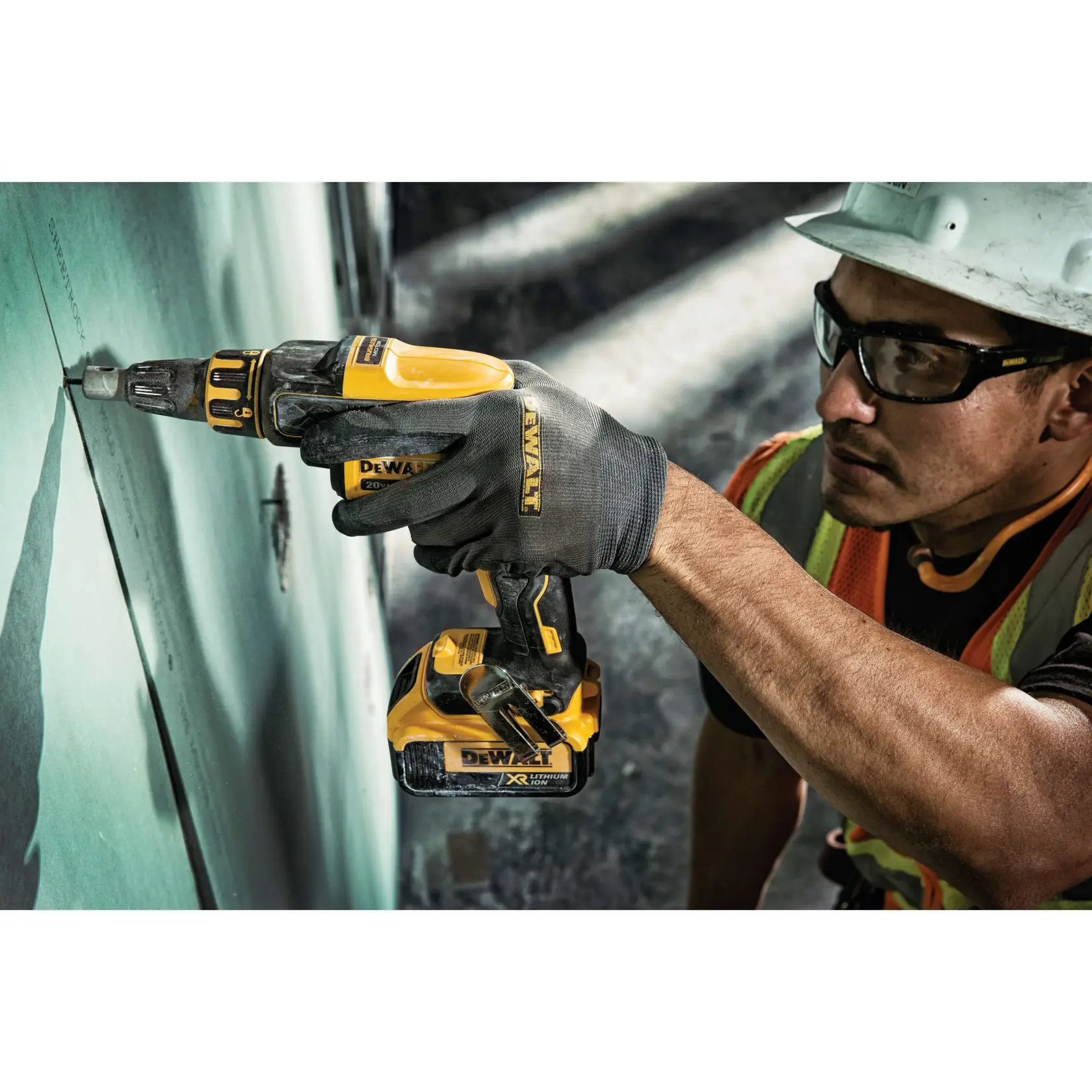 A person wearing safety gear uses a yellow DEWALT cordless power drill to secure a screw into a sheet of drywall.