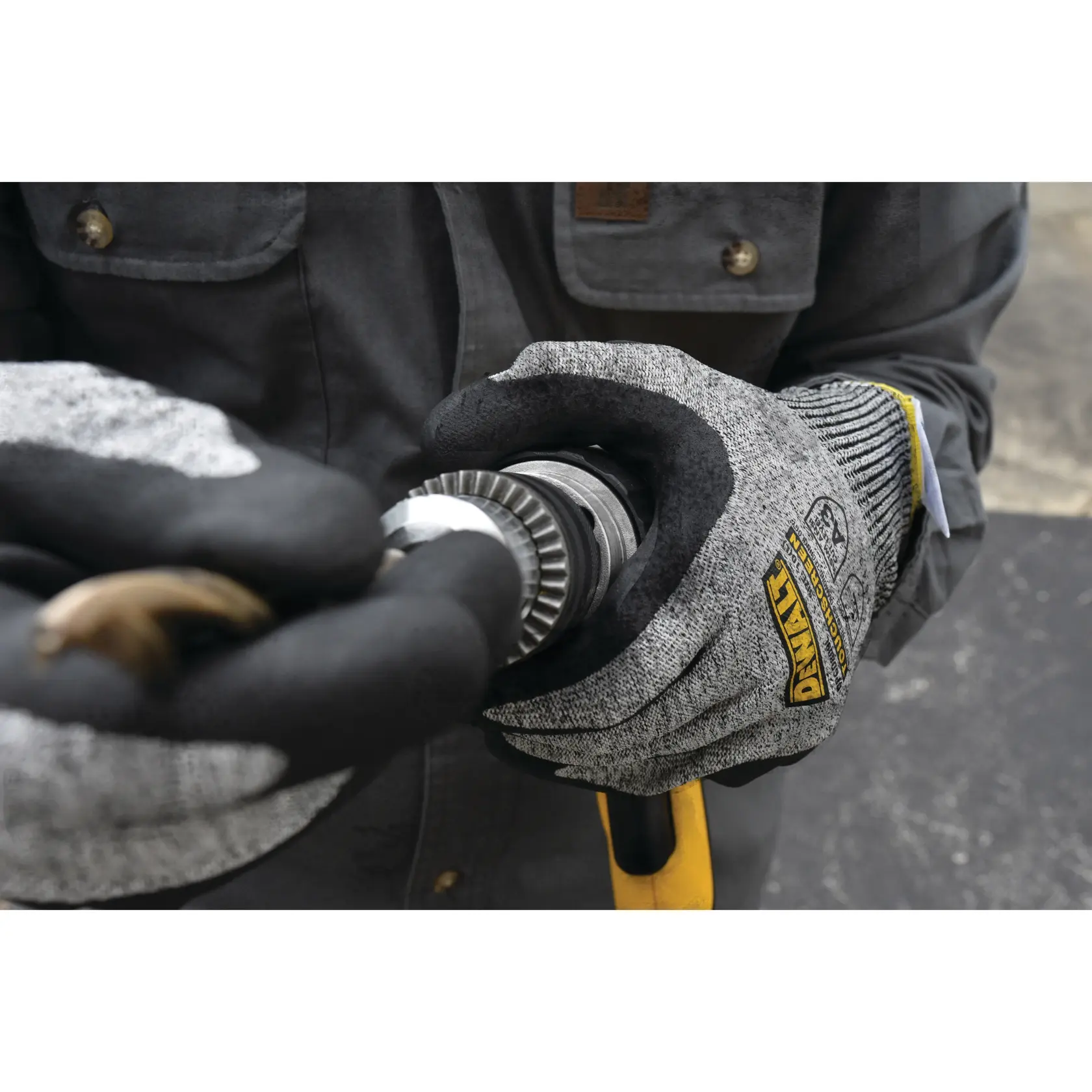 A close-up view of a person wearing gray and black DEWALT work gloves while holding a metal tool, with a dark jacket visible in the background.
