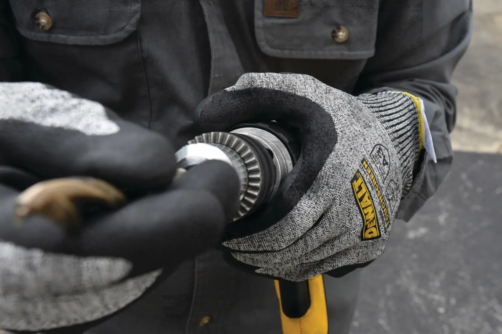 A person wearing DEWALT gray and black protective work gloves is holding a metal tool part, possibly for industrial or construction use.