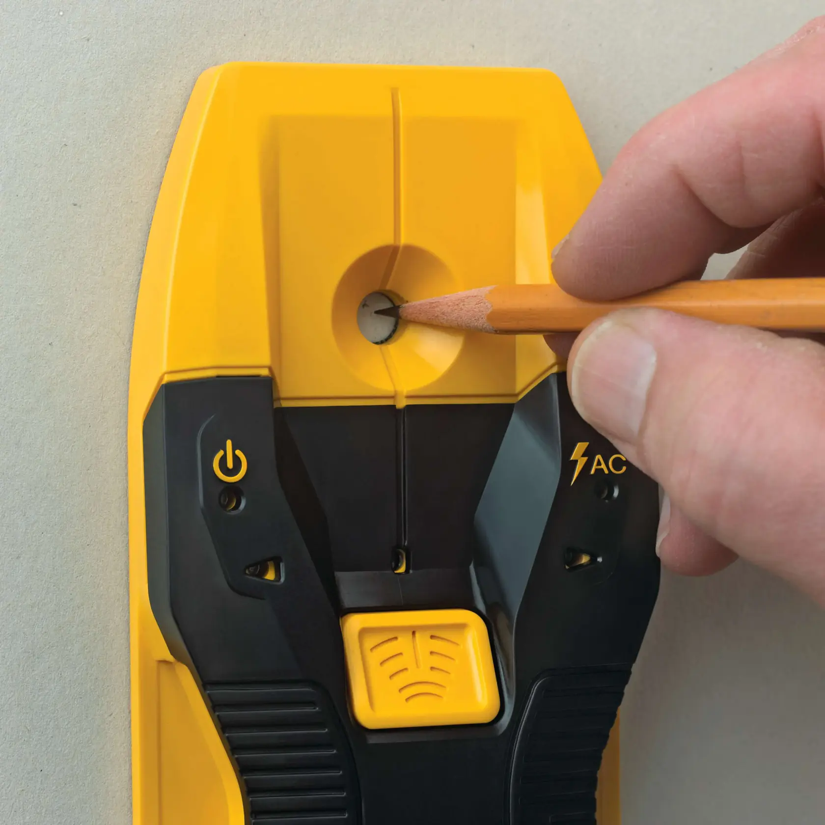 A close-up of a hand marking a spot on a wall using a pencil through the marking hole of a yellow and black DEWALT electronic stud finder.