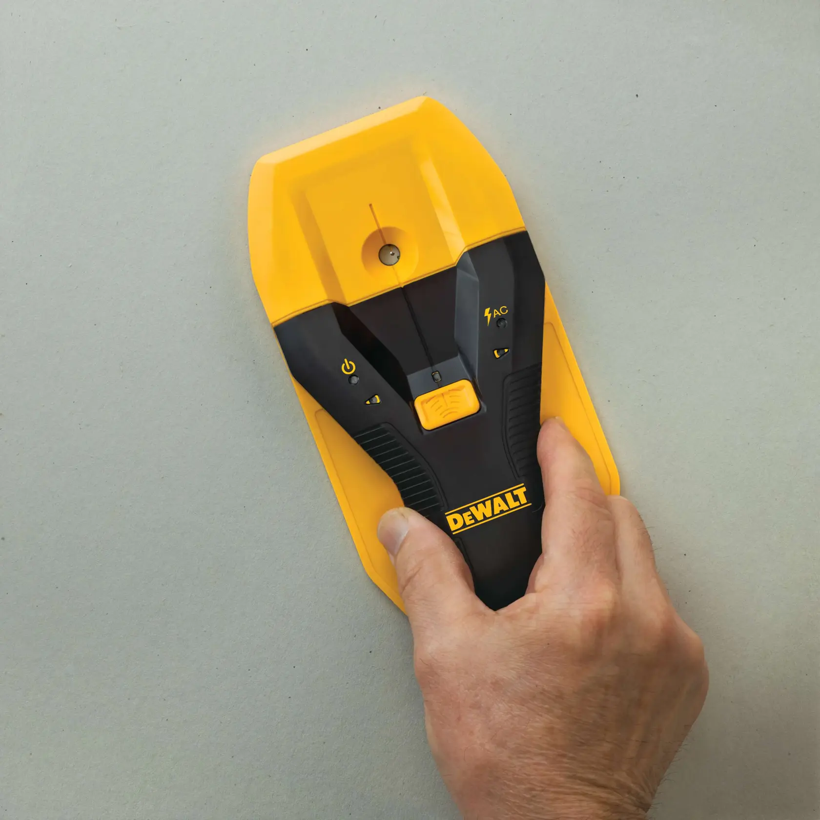 A hand holding a DEWALT stud finder device against a light gray wall. The device is black and yellow with visible control buttons.