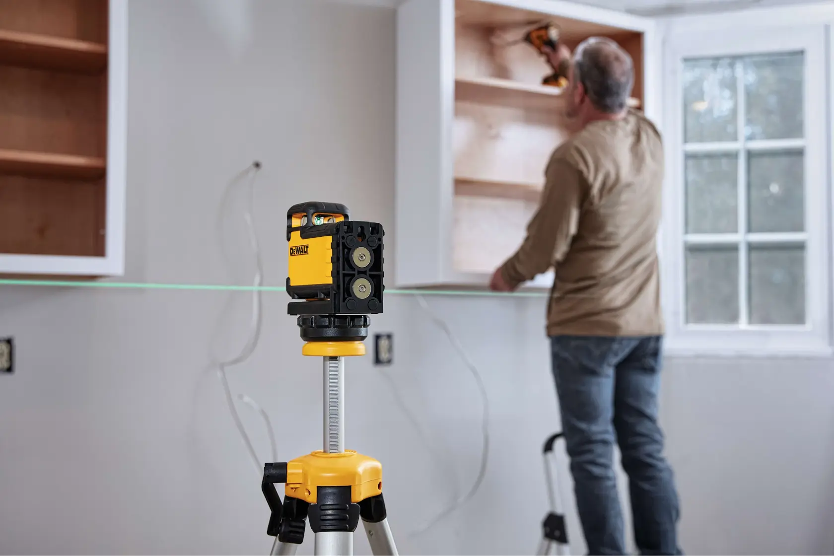 A DEWALT laser level mounted on a tripod is positioned in the foreground while a person installs wooden cabinets on a wall in a room. The laser line helps ensure precise alignment during installation.