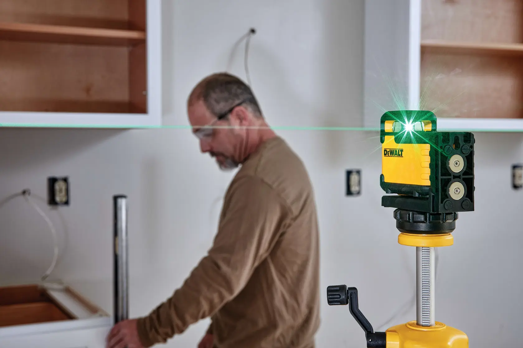 A DEWALT green laser level device is set up on a tripod in a room under renovation, projecting a green line across the wall. A person with a blurred face is seen in the background near a cabinet and work area.