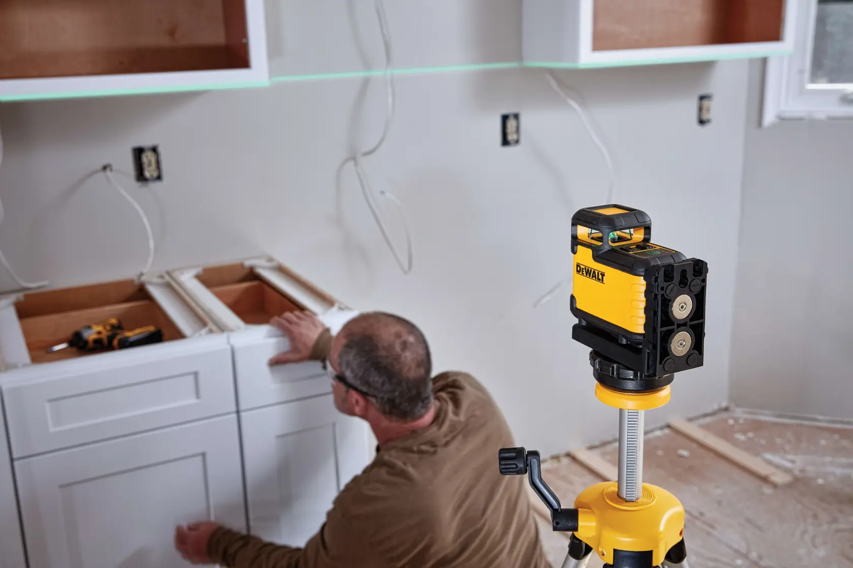 A DEWALT green cross line laser level (model DW03601CG) mounted on a tripod is being used in a kitchen installation. A person is aligning white cabinet drawers, with tools and wiring visible on the wall and countertops.