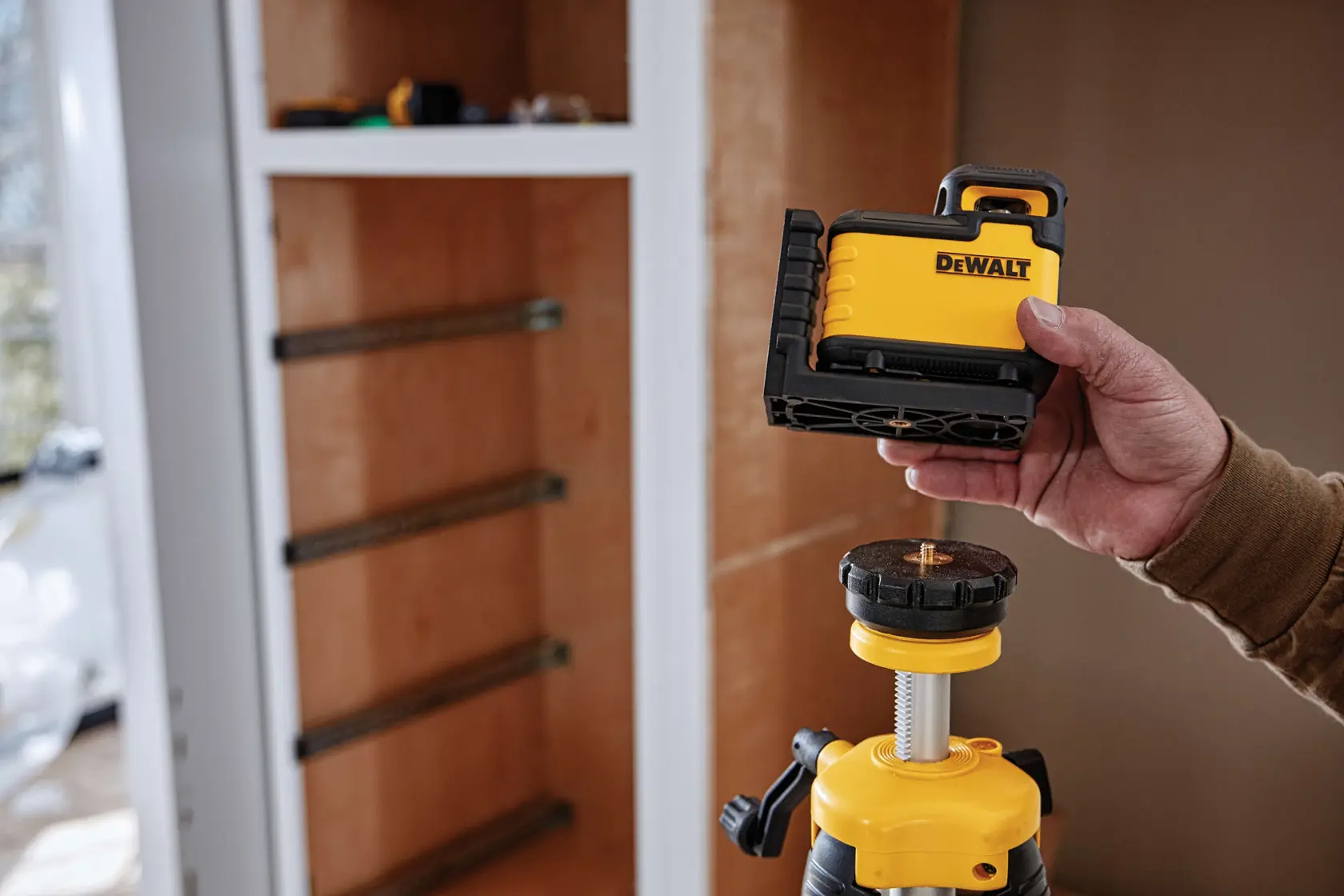A person holds a DEWALT laser level tool above a tripod in front of a wooden cabinet under construction.