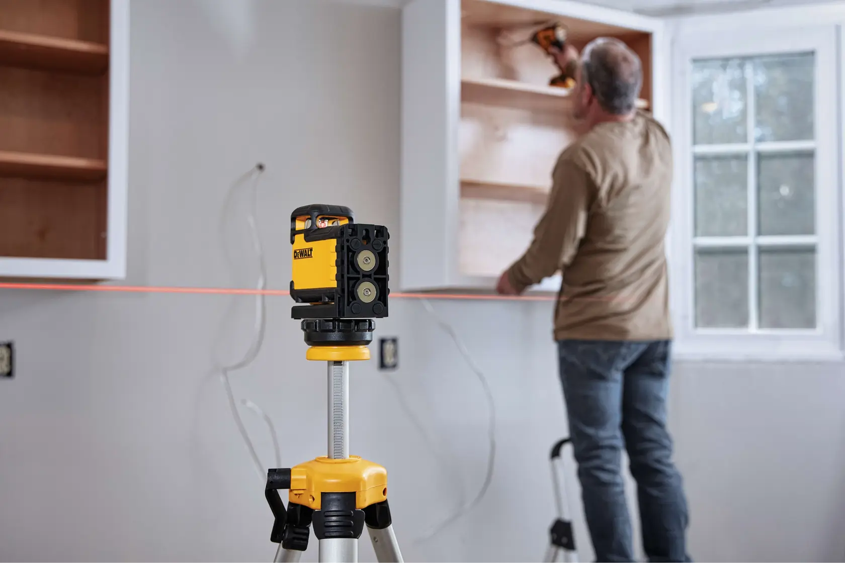 A DEWALT laser level mounted on a tripod projects a red line across a wall, aiding a person in installing cabinets in a room with a window.