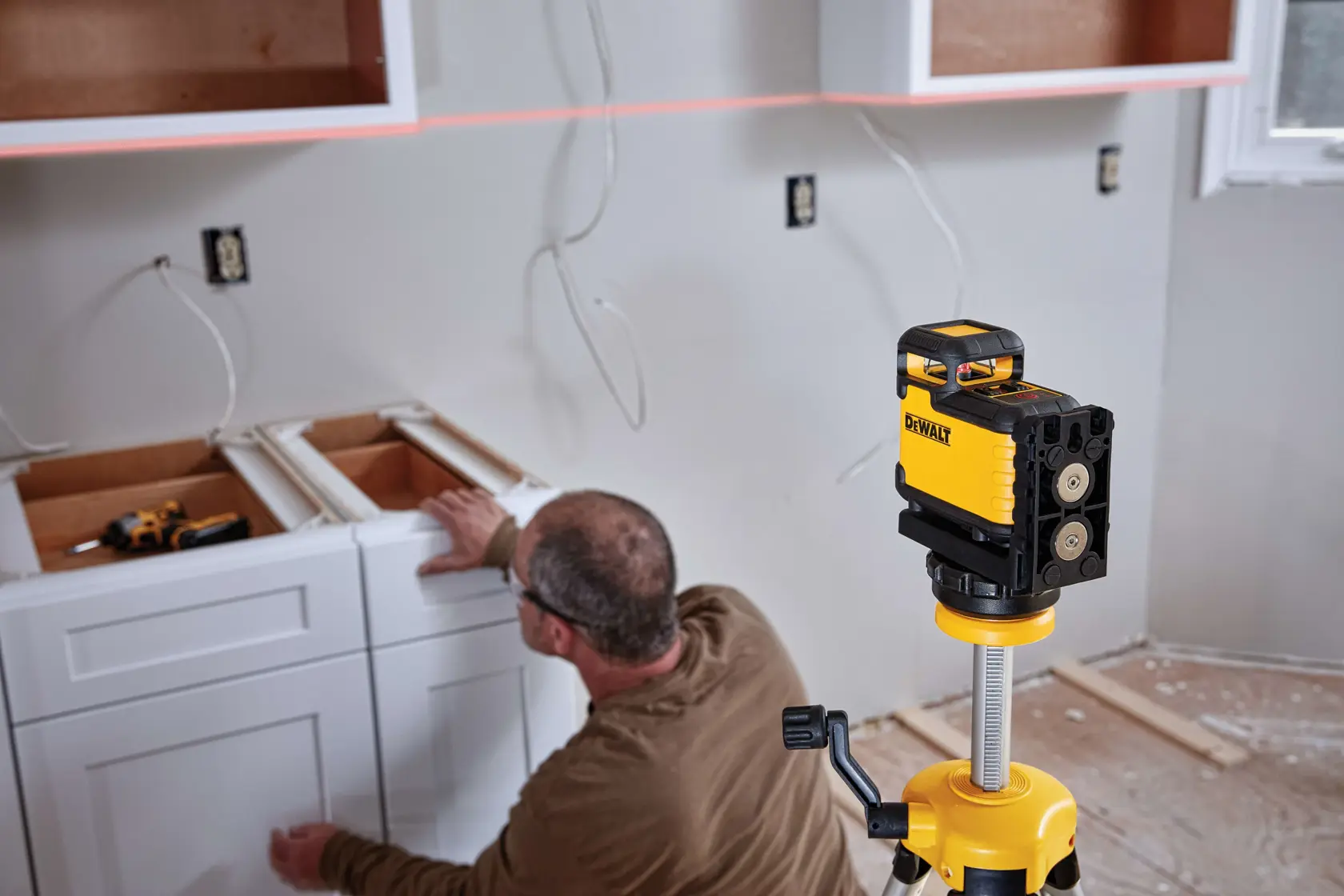A DEWALT laser level device is set up on a tripod in a kitchen under construction. A person is working on installing white cabinetry, and the laser projects a straight line across the wall for alignment.