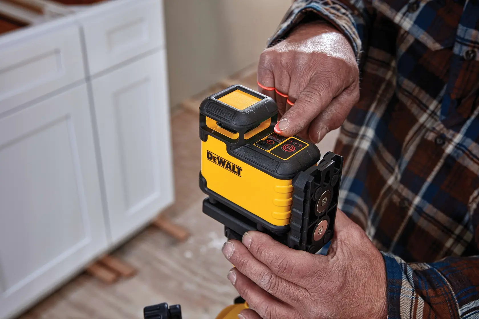 A person operating a DEWALT yellow and black laser level device, pressing a button on top. The device is mounted on a tripod, and is used for precise leveling and alignment in construction or renovation. White cabinetry and a wooden floor are visible in the background.