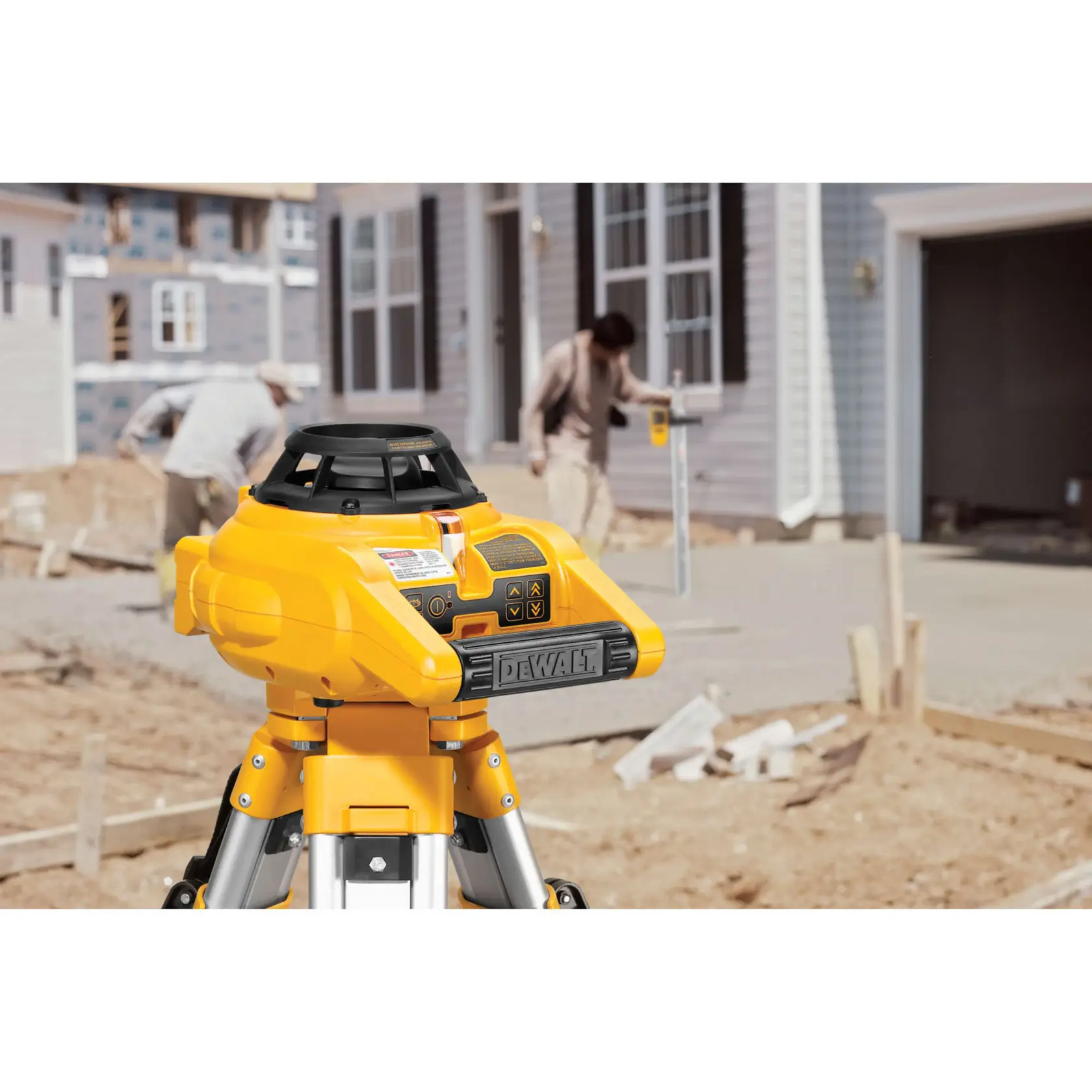 A DEWALT laser level device mounted on a tripod is positioned at a construction site in front of a house, with two workers in the background performing leveling and measuring tasks.