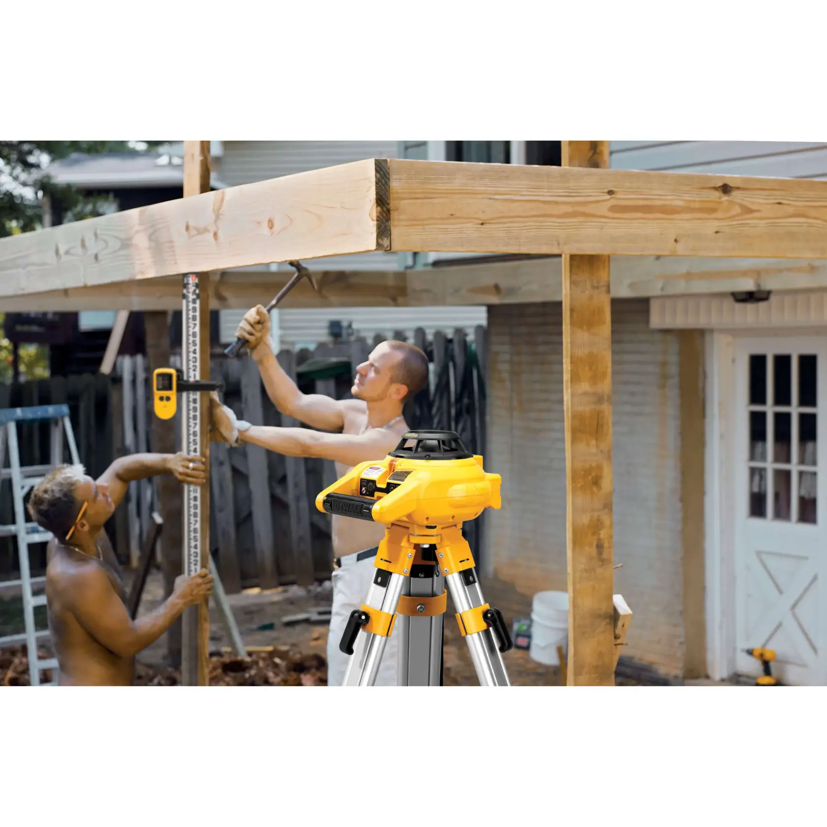 A DEWALT DW074KD rotary laser level is set up on a tripod in the foreground of a construction scene. Two people are working on wooden beams, one holding a measuring rod with a laser detector, and the other using a hammer. The setting appears to be outdoors near a house.