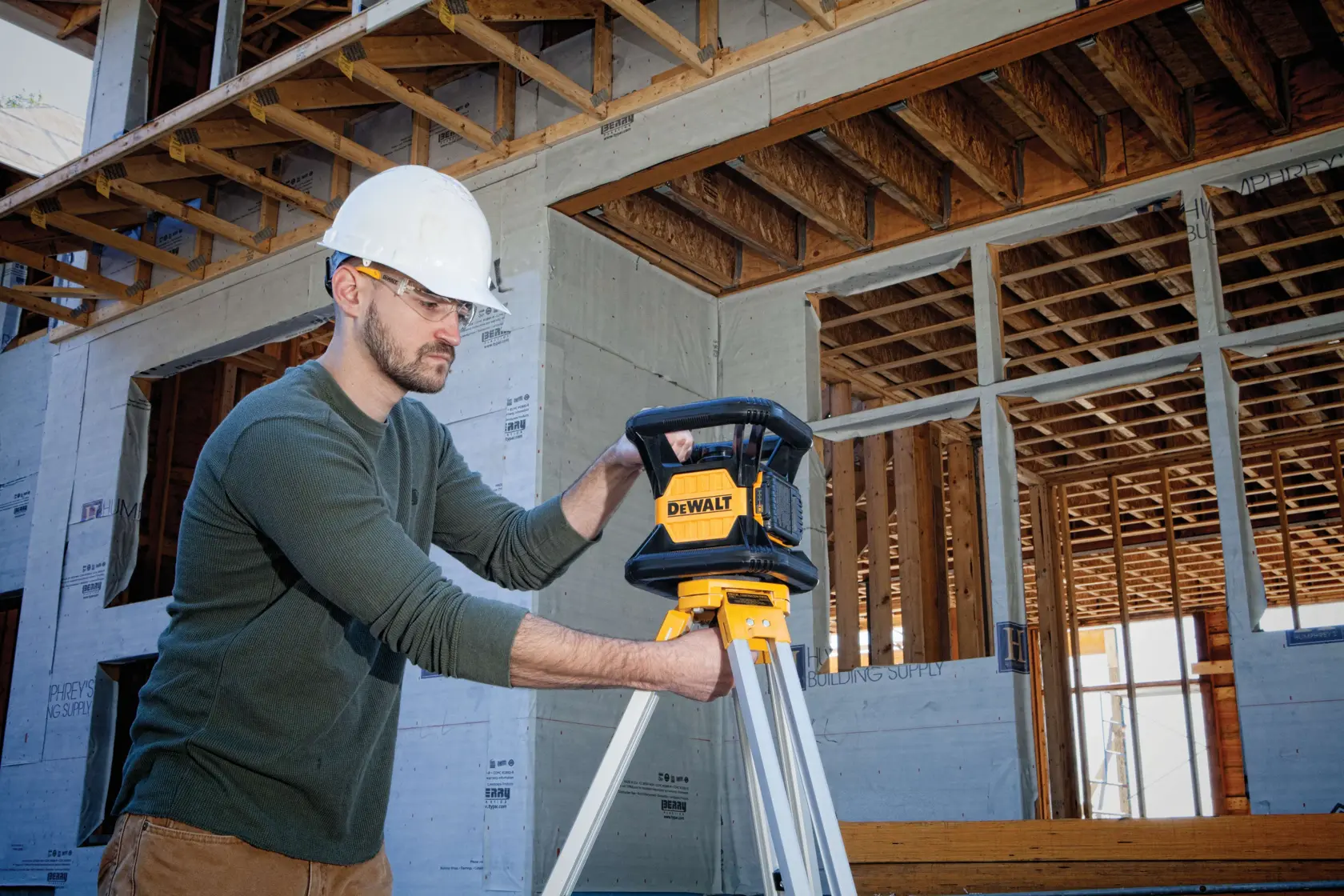A person wearing a white hard hat is adjusting a DEWALT DW079LG laser level on a tripod at a construction site with wooden framing and partially built walls.