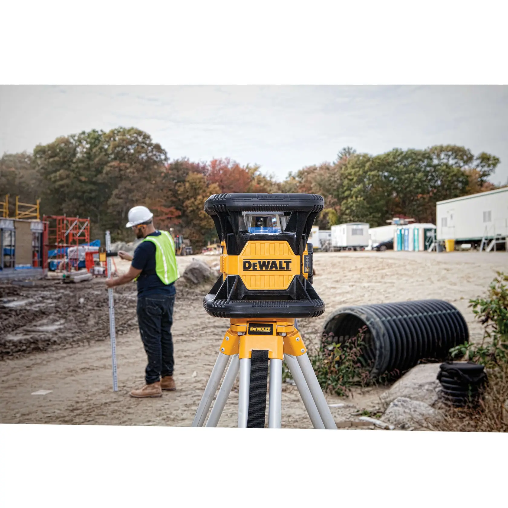 A DEWALT DW079LR rotary laser level mounted on a tripod is shown at a construction site. In the background, a worker wearing a safety vest and helmet is measuring with a leveling rod, and there are portable buildings and construction materials visible.