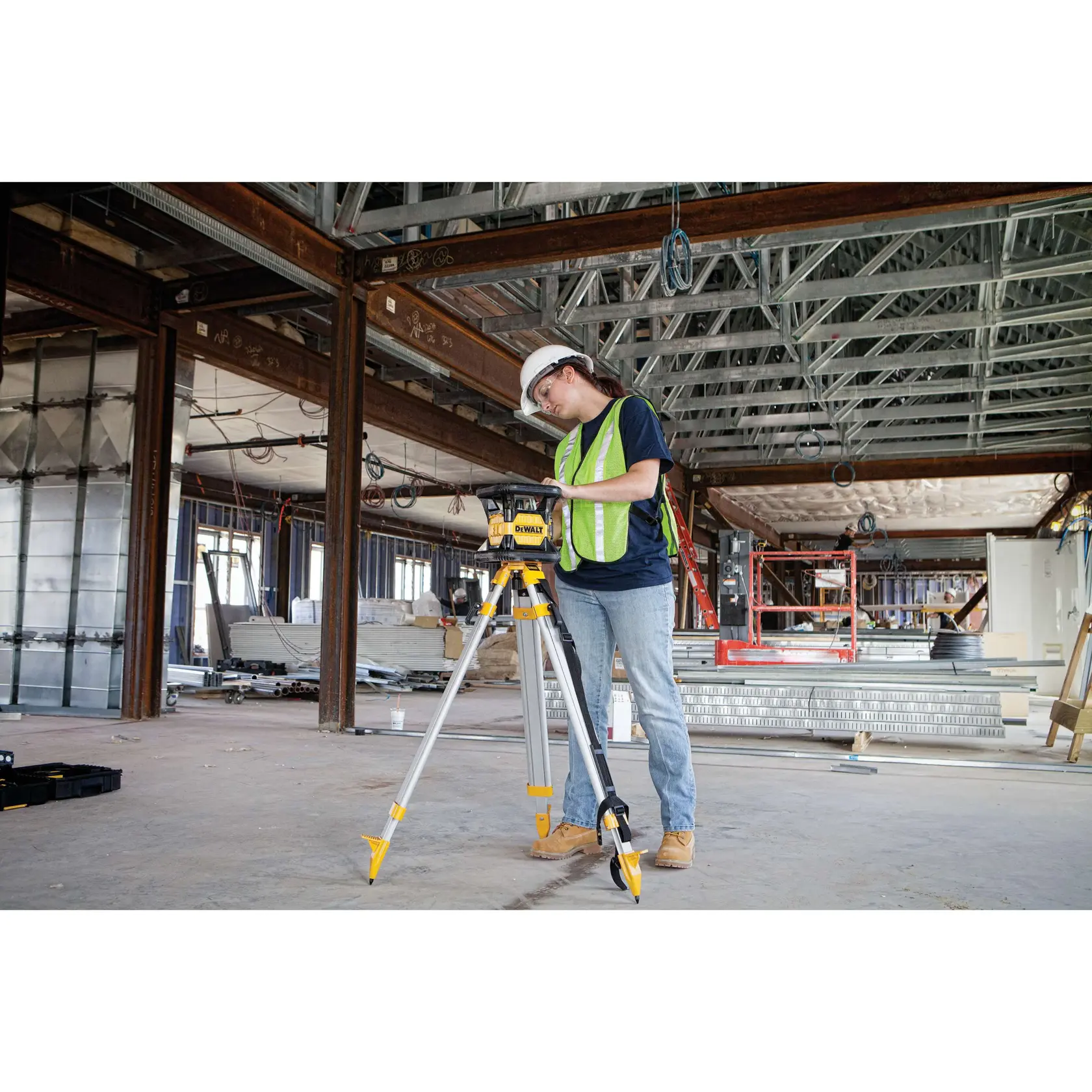 A person wearing a safety helmet and a reflective vest operates a DEWALT DW079LR rotary laser level mounted on a tripod at a construction site with exposed steel beams and building materials.