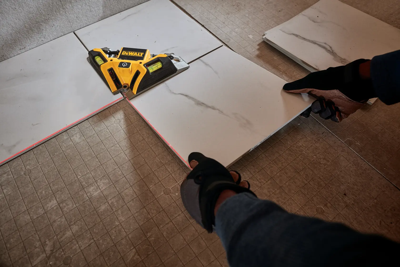 A DEWALT laser level tool is being used to align ceramic tiles on a floor. The tool emits a red laser line to help with precise tile placement, and gloved hands are adjusting the tiles.