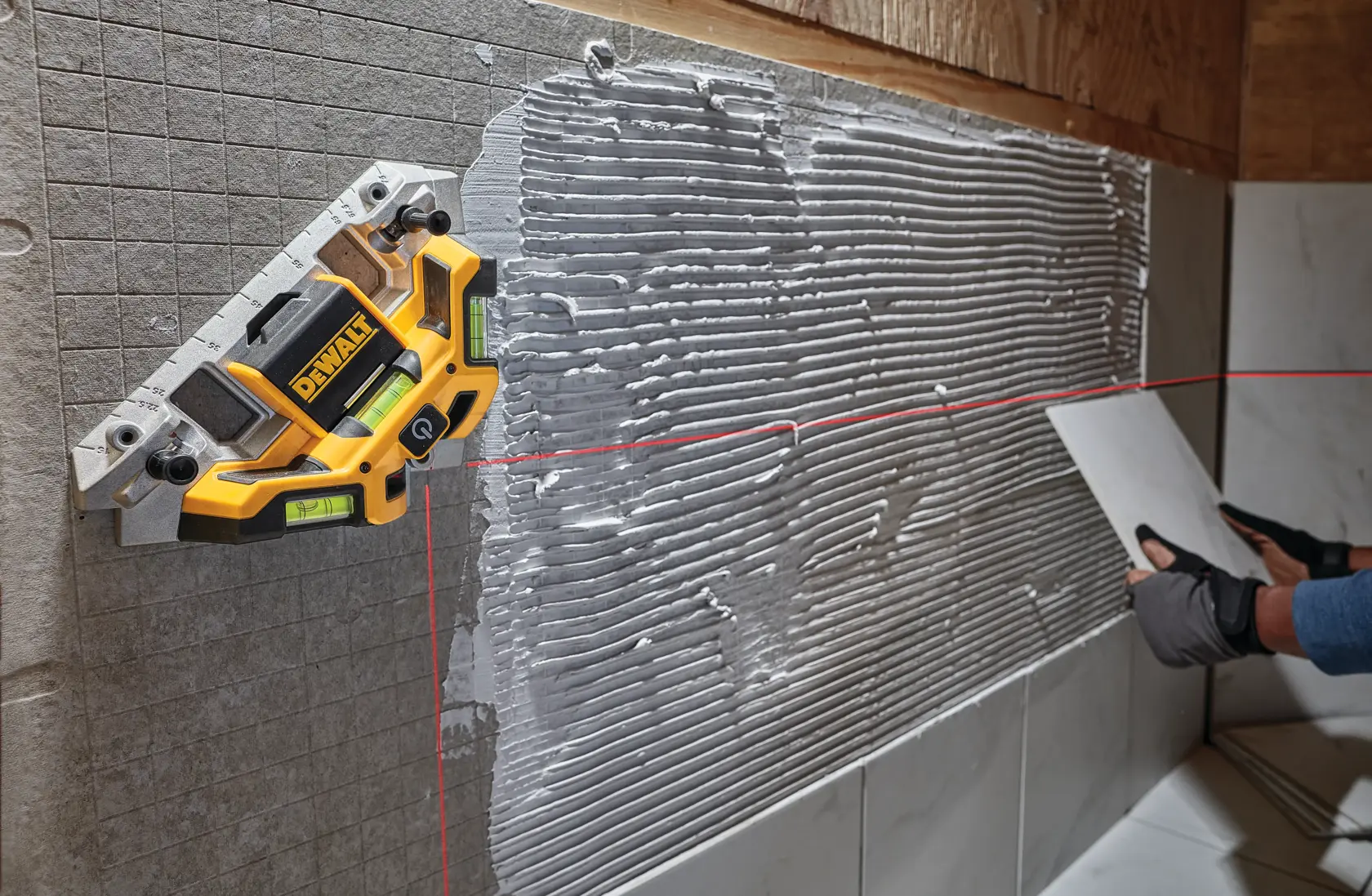 A DEWALT laser level is mounted on a wall to project a horizontal and vertical red laser line, assisting in the precise installation of tiles. The wall is partially covered with adhesive and tiles, and a person wearing gloves is positioning a tile according to the laser guide.