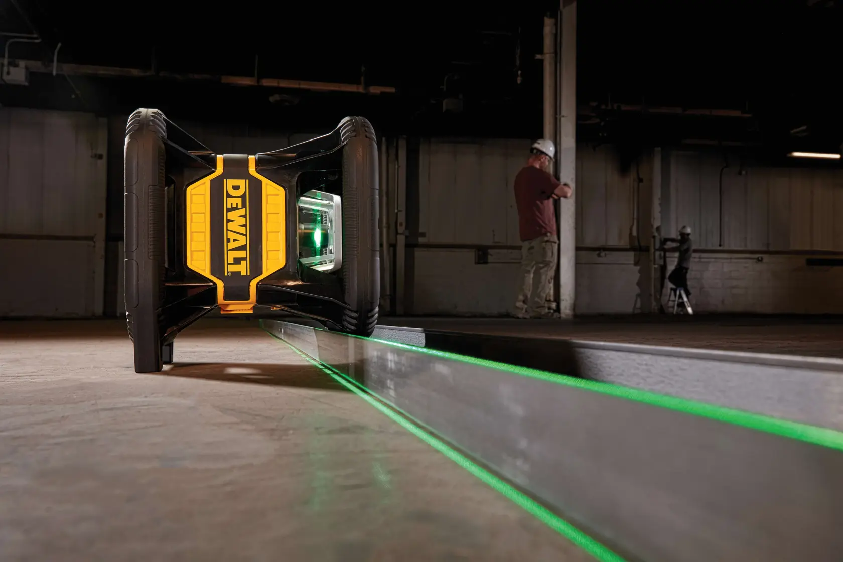A DEWALT green laser level device is positioned on the floor, projecting a bright green laser line across the surface. The setting appears to be an industrial or construction area, with two people wearing safety gear in the background.