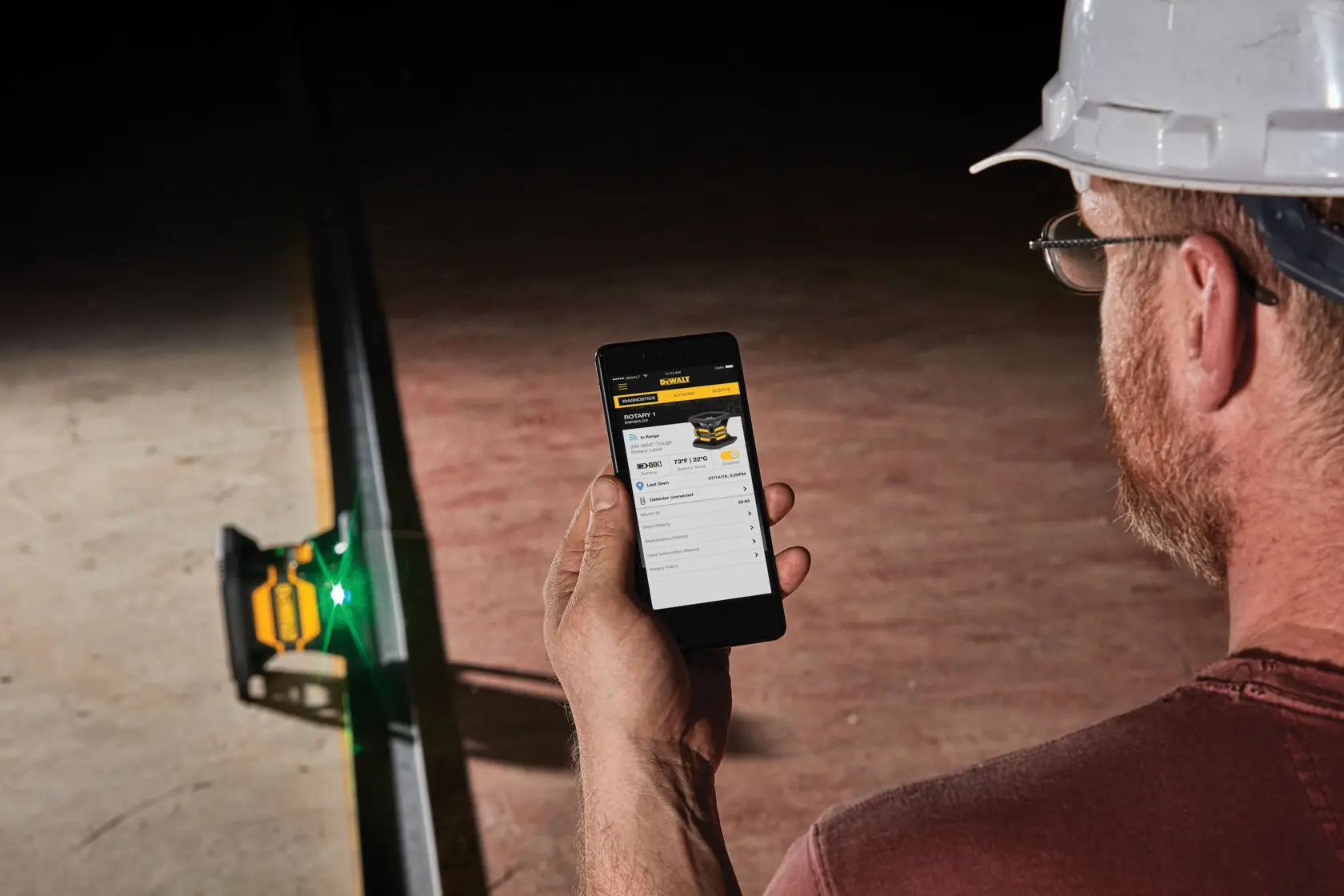 A person wearing a hard hat and glasses is using a smartphone to control or check settings for a DEWALT laser level device on a construction site.